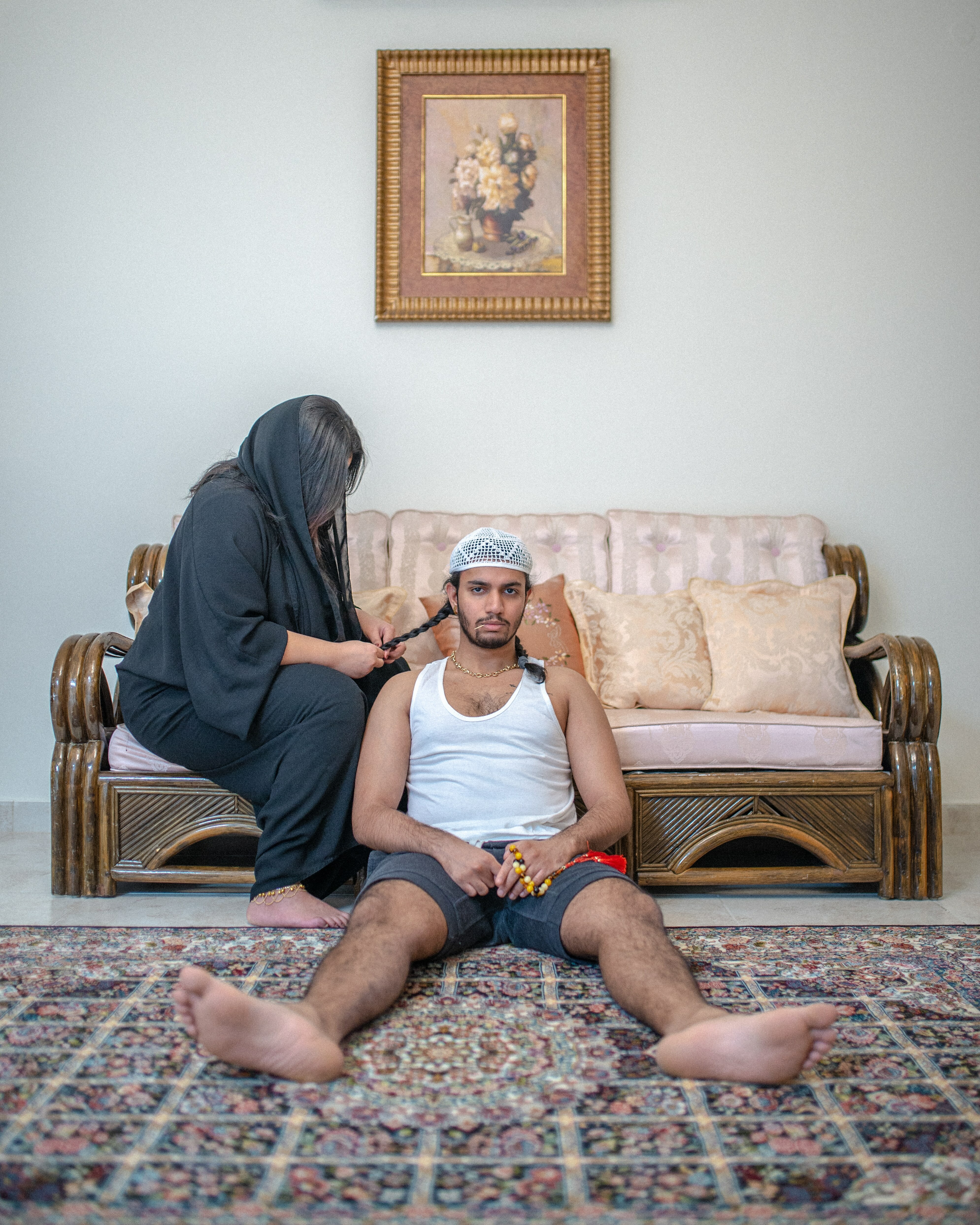 Framed floral painting on wall, wooden sofa, patterned rug, man and woman seated on floor.