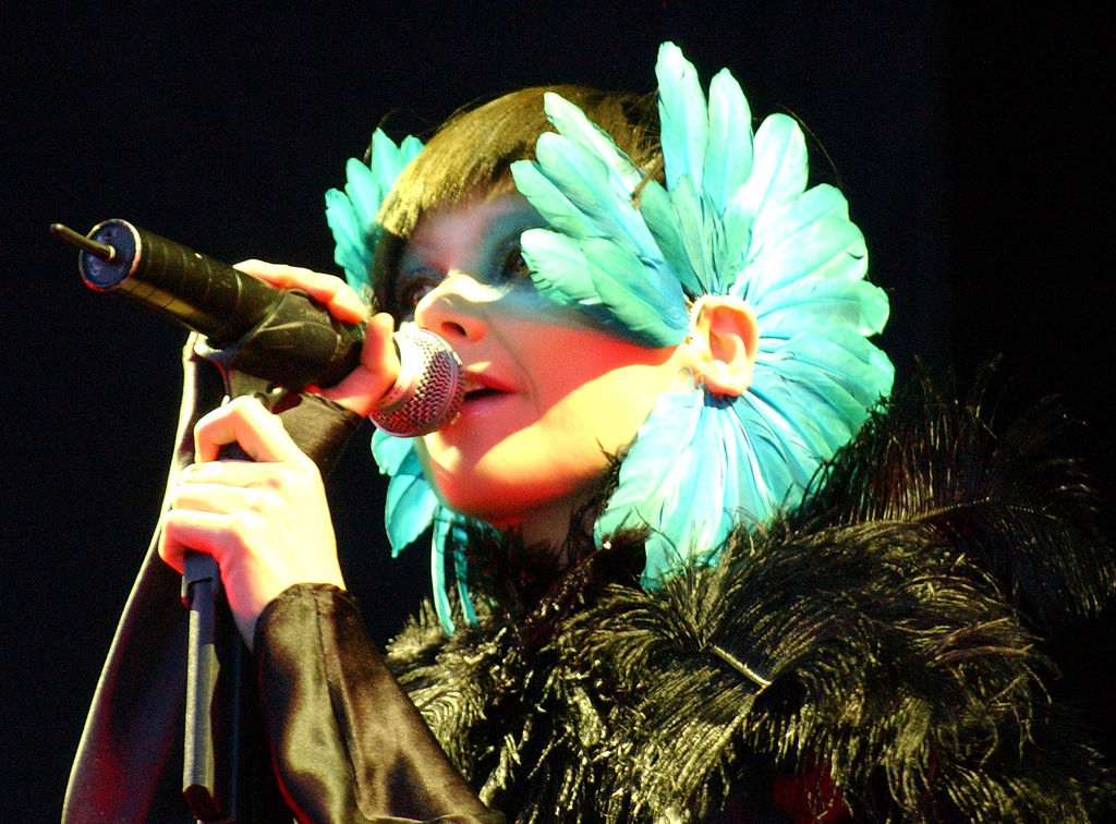 Performer singing into microphone wearing large turquoise feathered mask covering half their face and dark feathered costume.