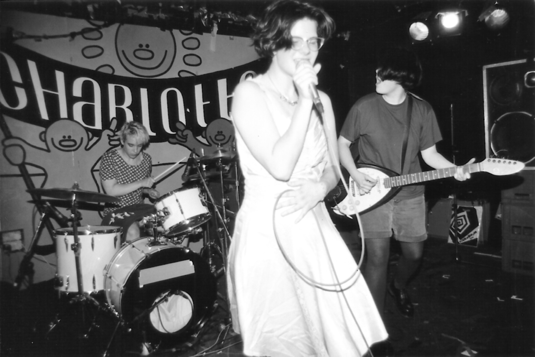 A black and white image of a band performing on stage, with the singer in a white dress and the guitarist and drummer visible.