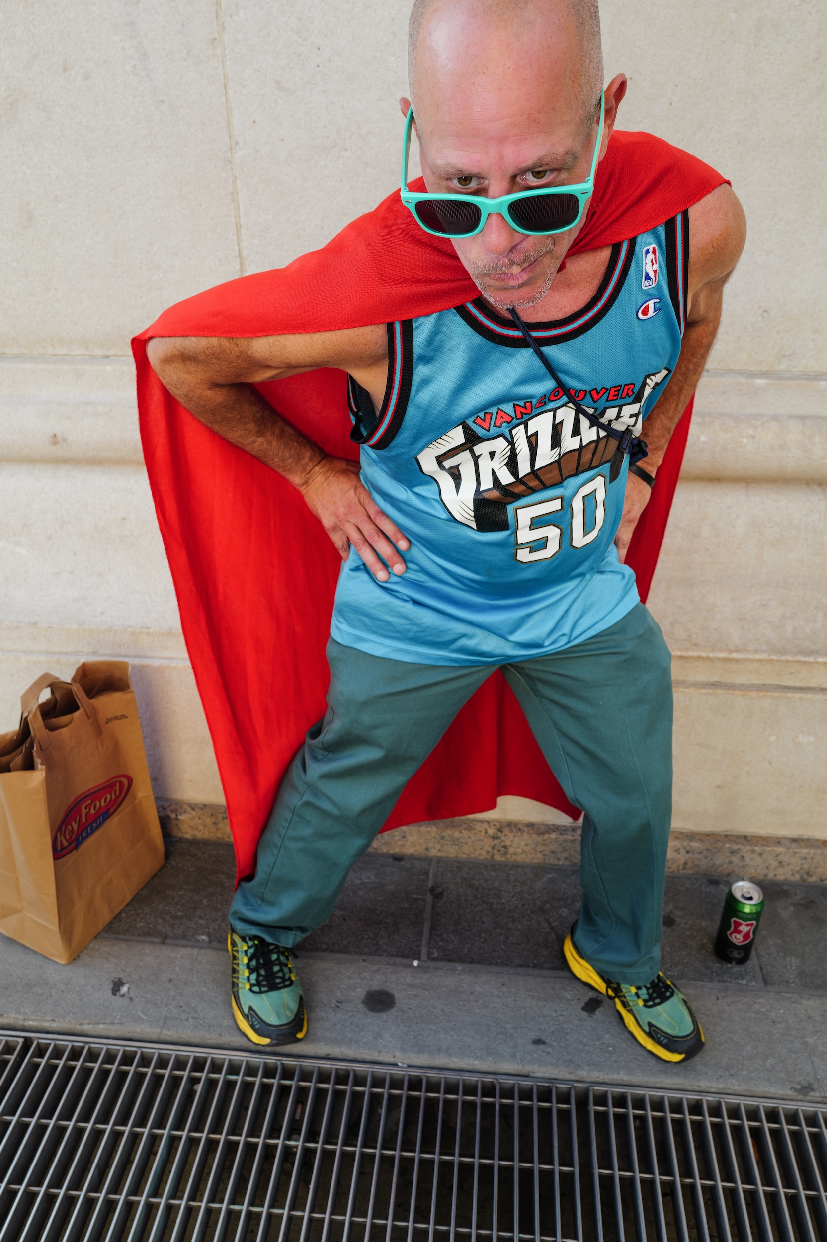 Bald man wearing red cape, blue Grizzlies basketball jersey, green sunglasses and trousers, posing with hand on hip on concrete steps.
