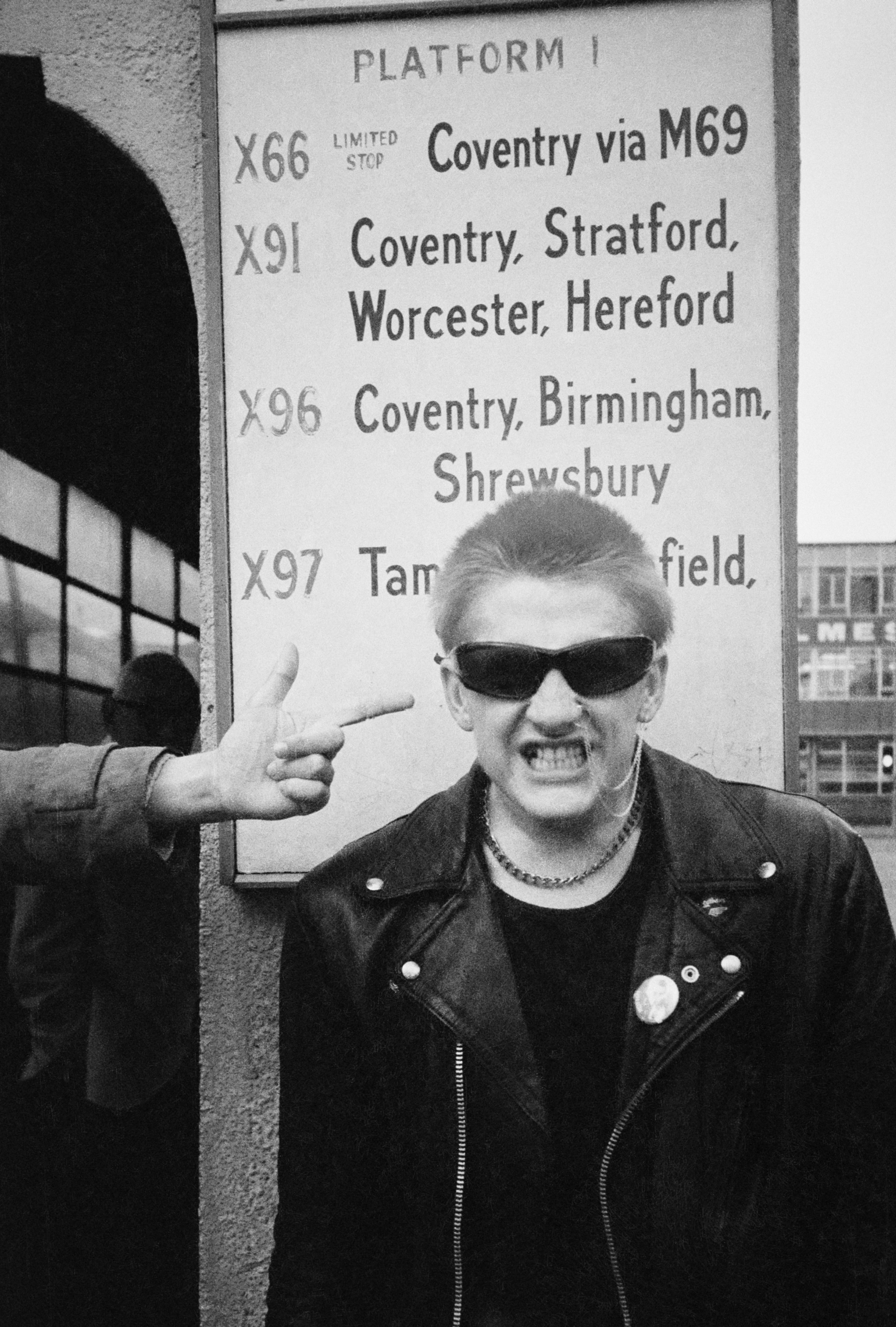 Man in leather jacket and sunglasses making finger gun gesture at temple beside railway platform sign listing bus routes to Coventry.