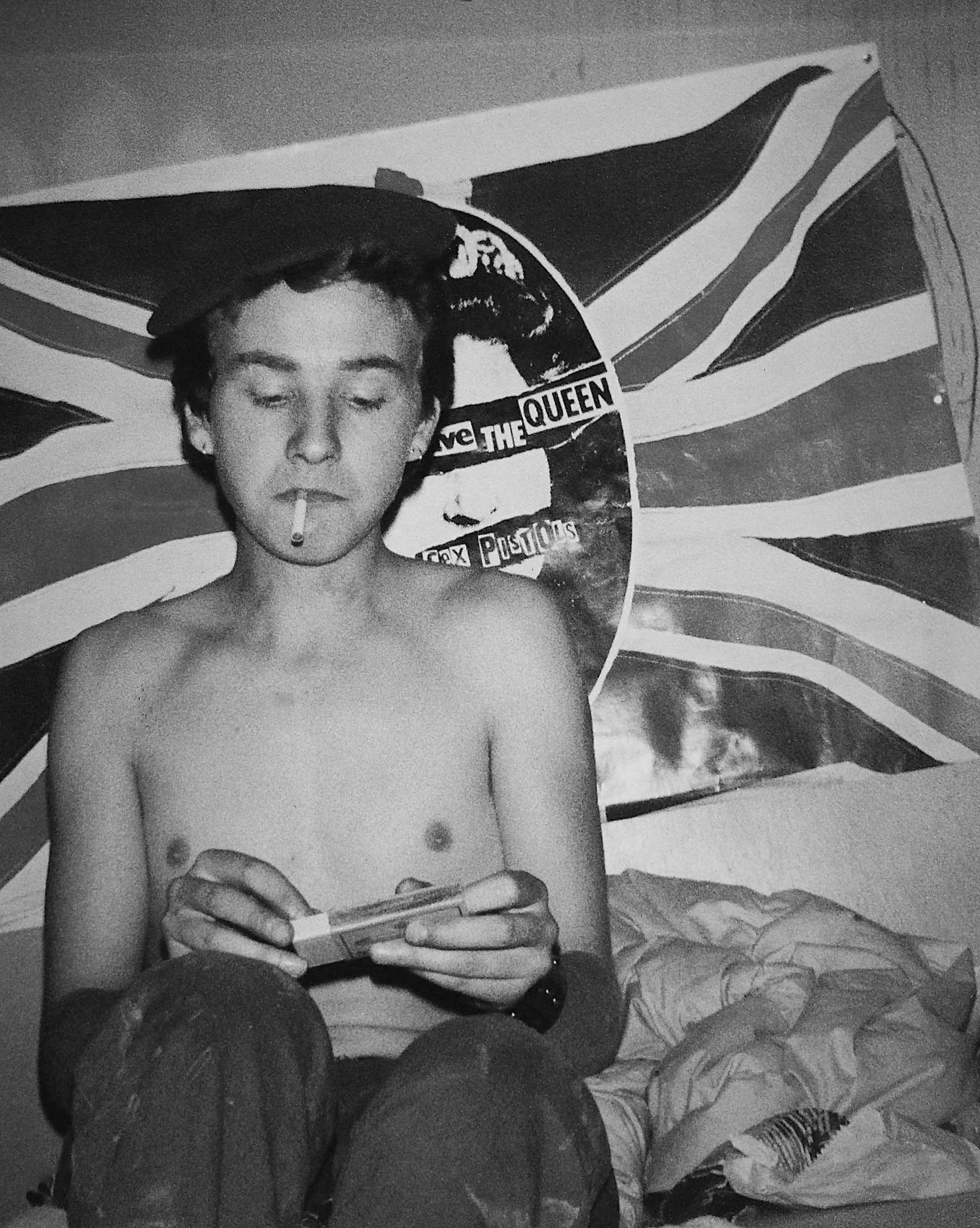 Shirtless young man with cigarette reading on bed, Union Jack flag and Sex Pistols "God Save the Queen" record visible behind him.