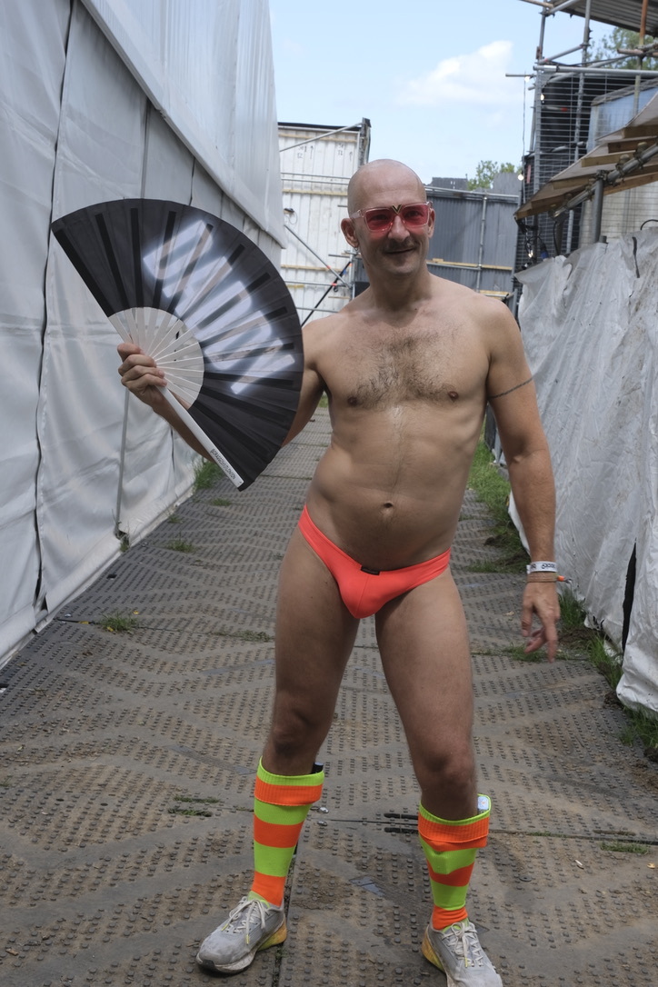 Shirtless man in orange briefs and striped socks holding black fan on industrial rooftop with concrete structures and scaffolding.
