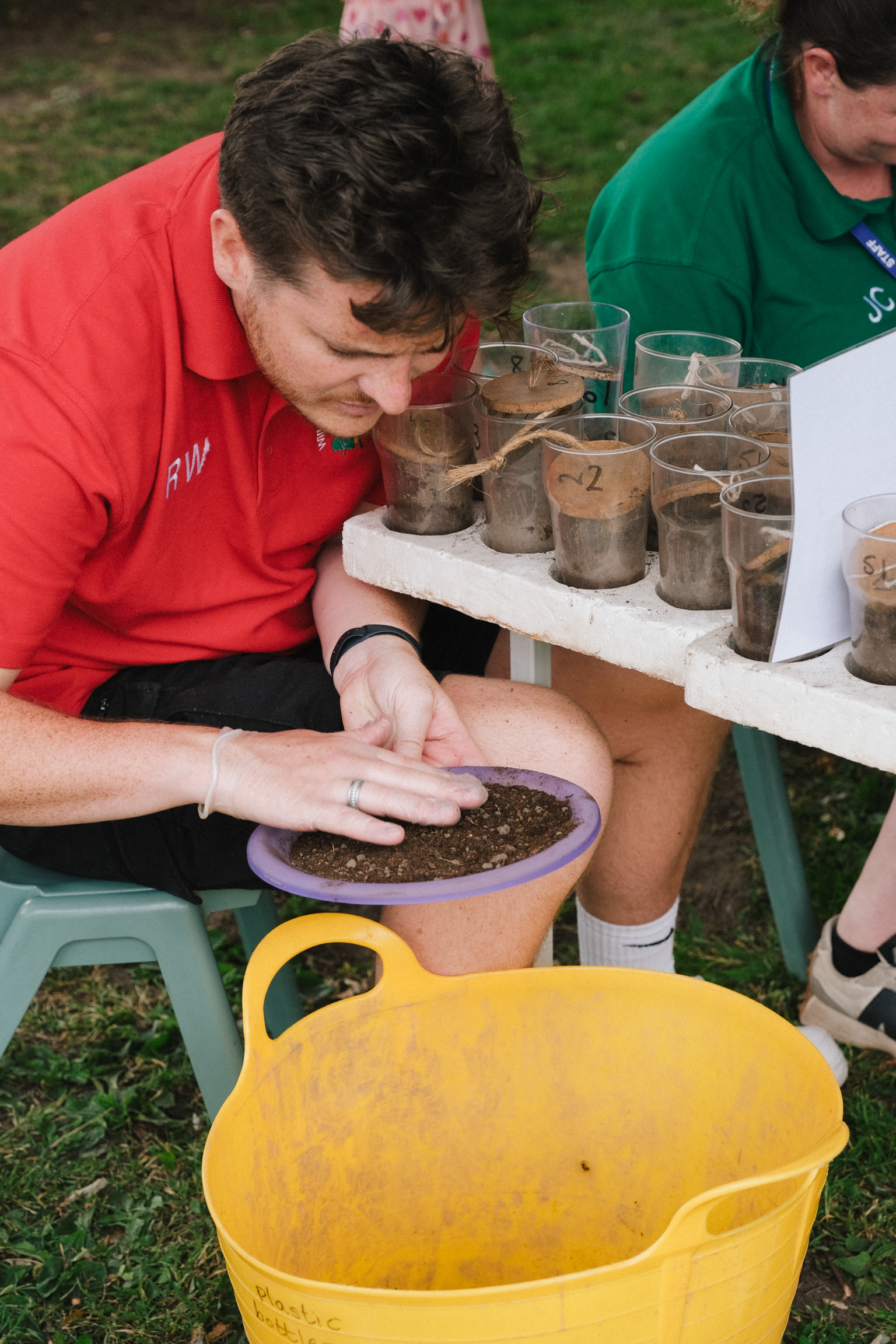 Man in red shirt examining soil samples in plastic containers on white table, with yellow bucket and various glass jars containing dirt.