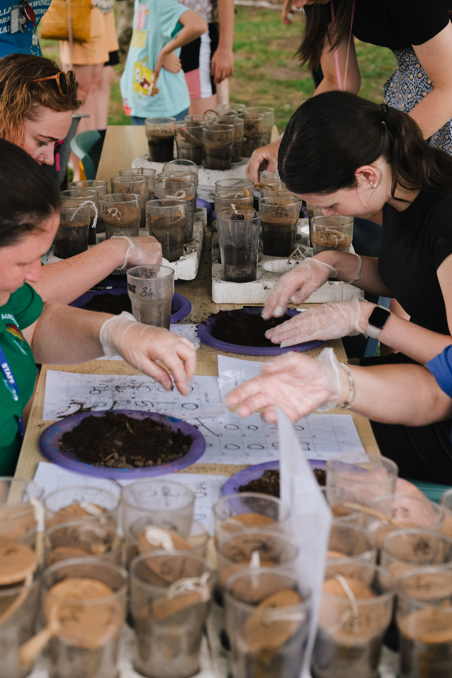 Children examining soil samples and sediment cores at outdoor educational activity with glass jars and worksheets on table.