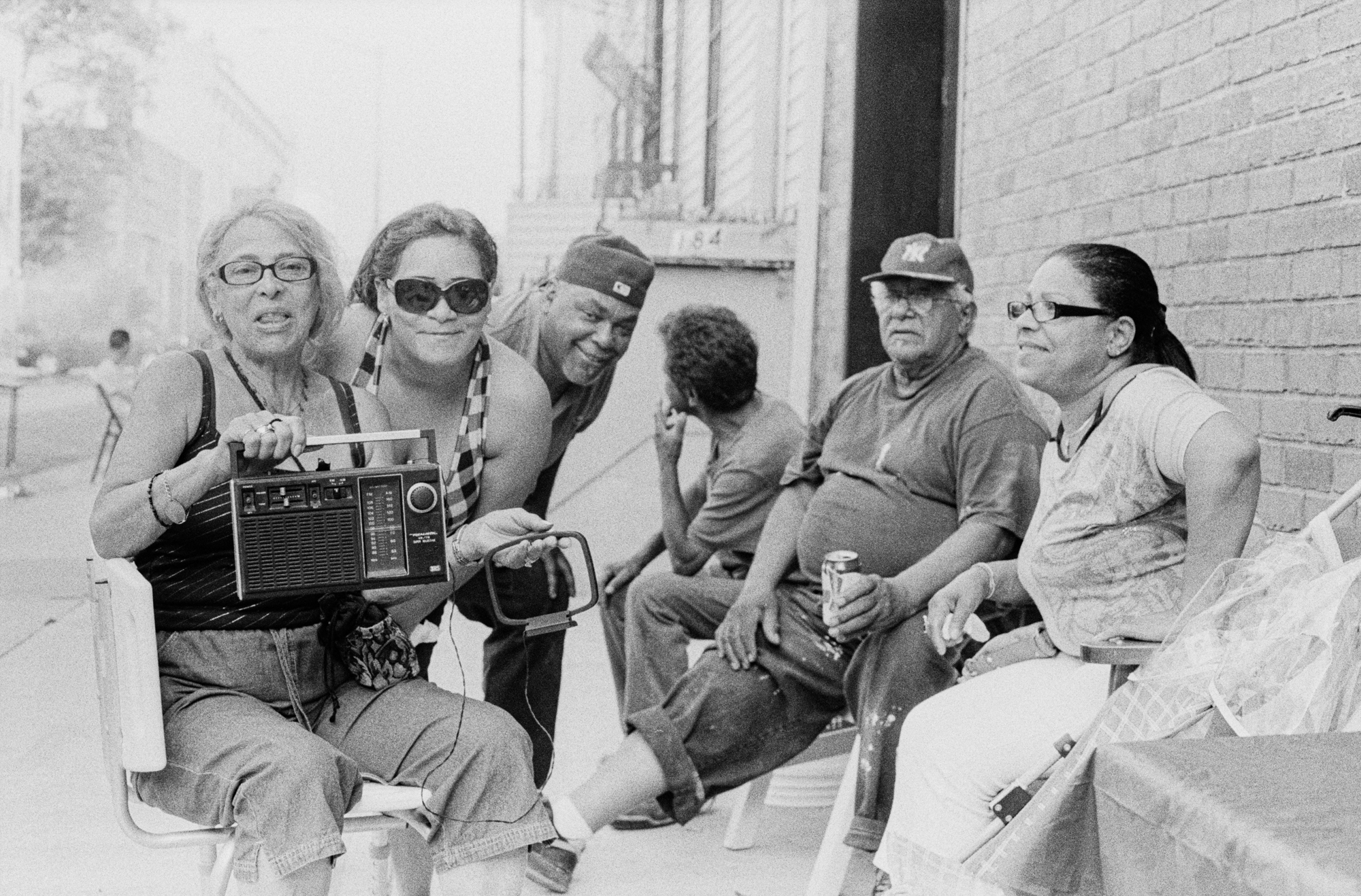 Group of 6 adults, some with glasses, hats and casual clothing, sitting on a wall outside a building.