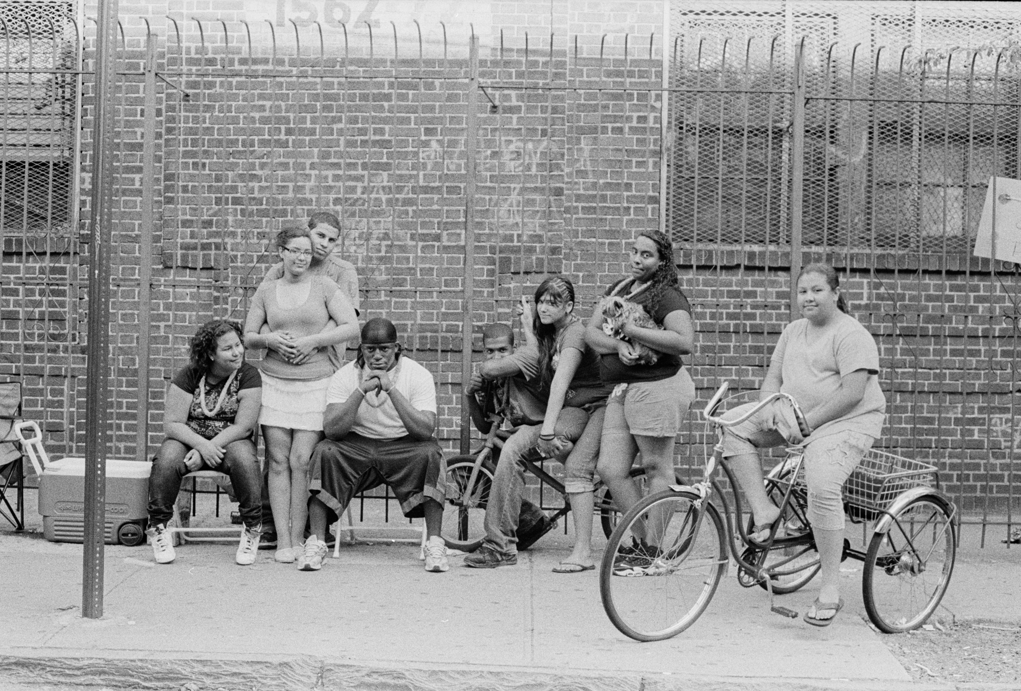 Group of women and children sitting and standing on a sidewalk by a brick wall.