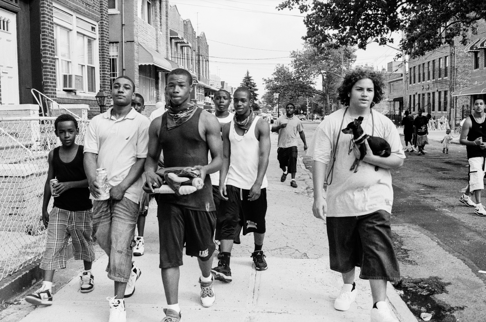 A group of young people walking down a city street, some wearing casual clothes like T-shirts and shorts, with a mix of genders and ethnicities visible.