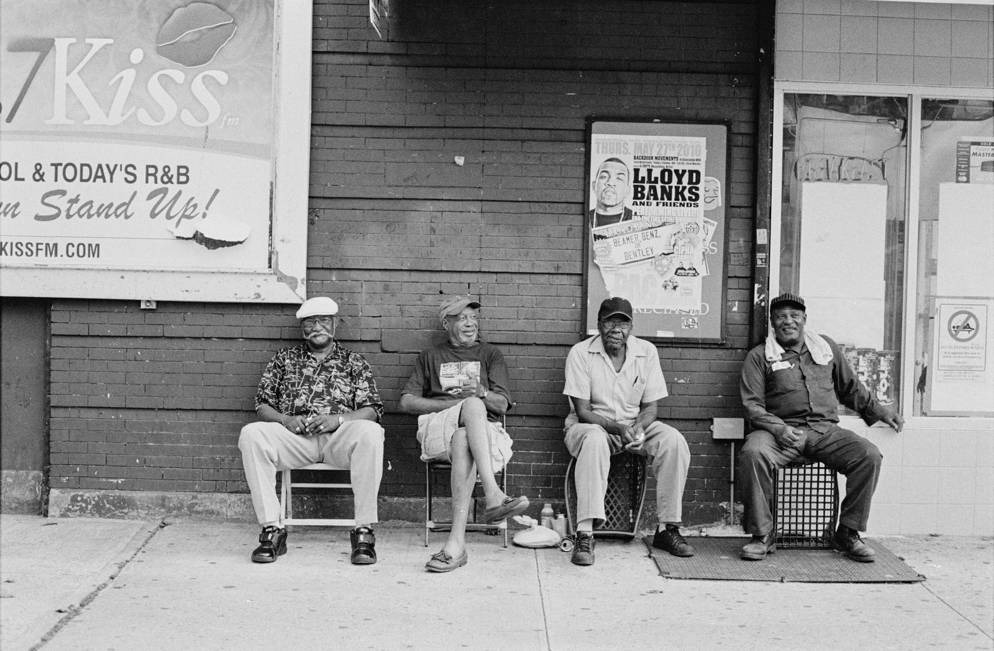 Older Black men seated on chairs outside a building with various signage and advertising.