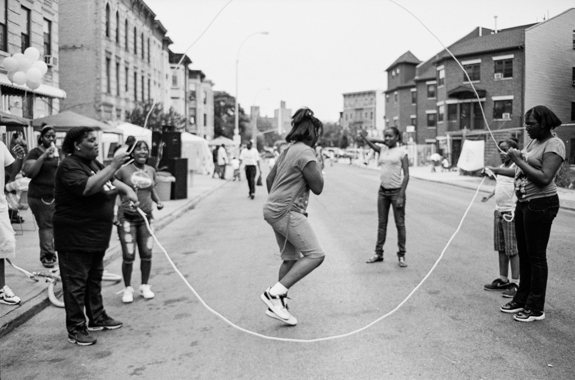 A black and white image of a busy urban street scene. In the centre, a person is jumping rope on the pavement, surrounded by other people watching. Buildings, shops, and traffic can be seen in the background.