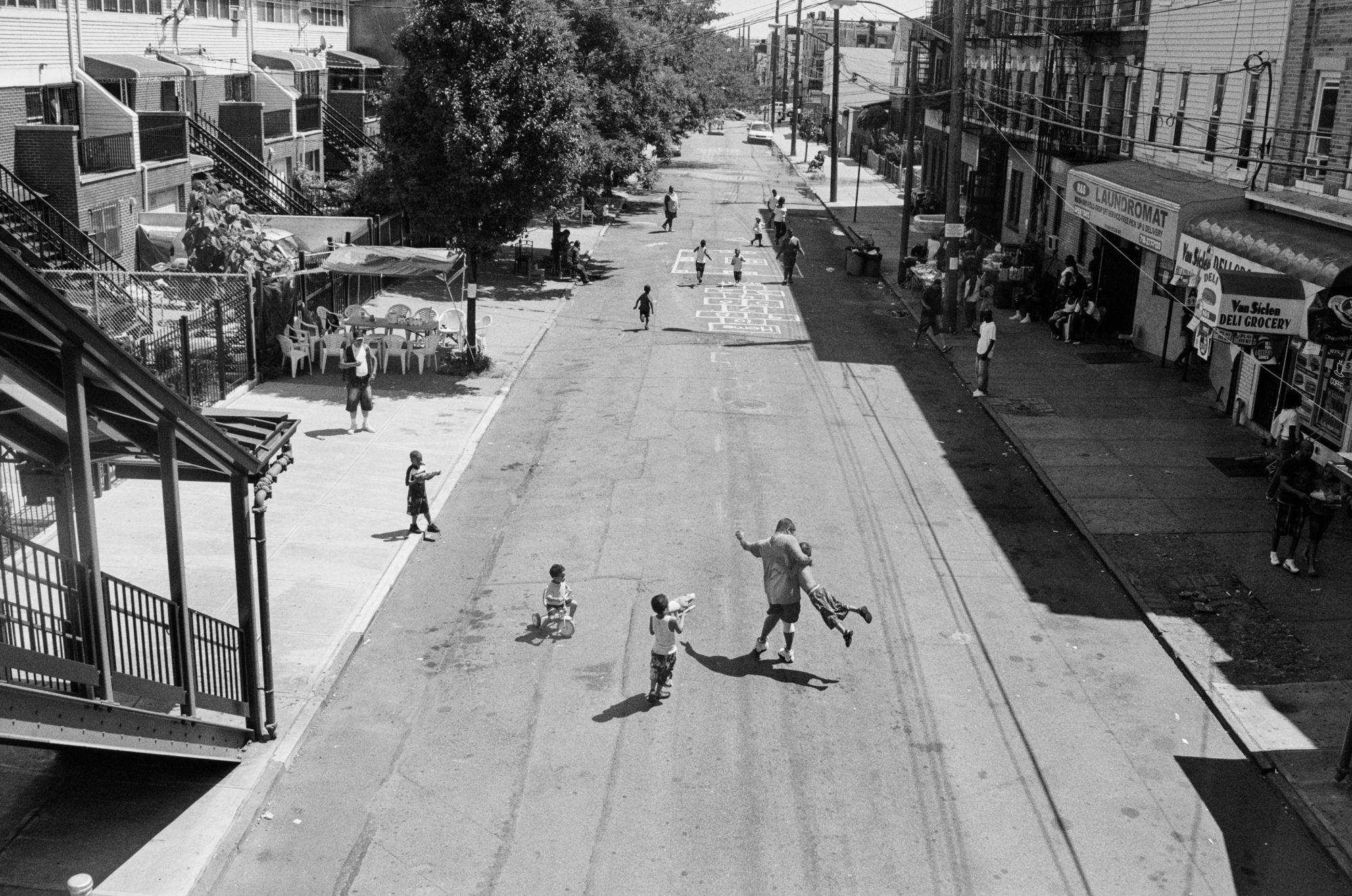 Busy street scene with people walking, riding bikes, and playing on the pavement. Storefronts and buildings line both sides of the road.