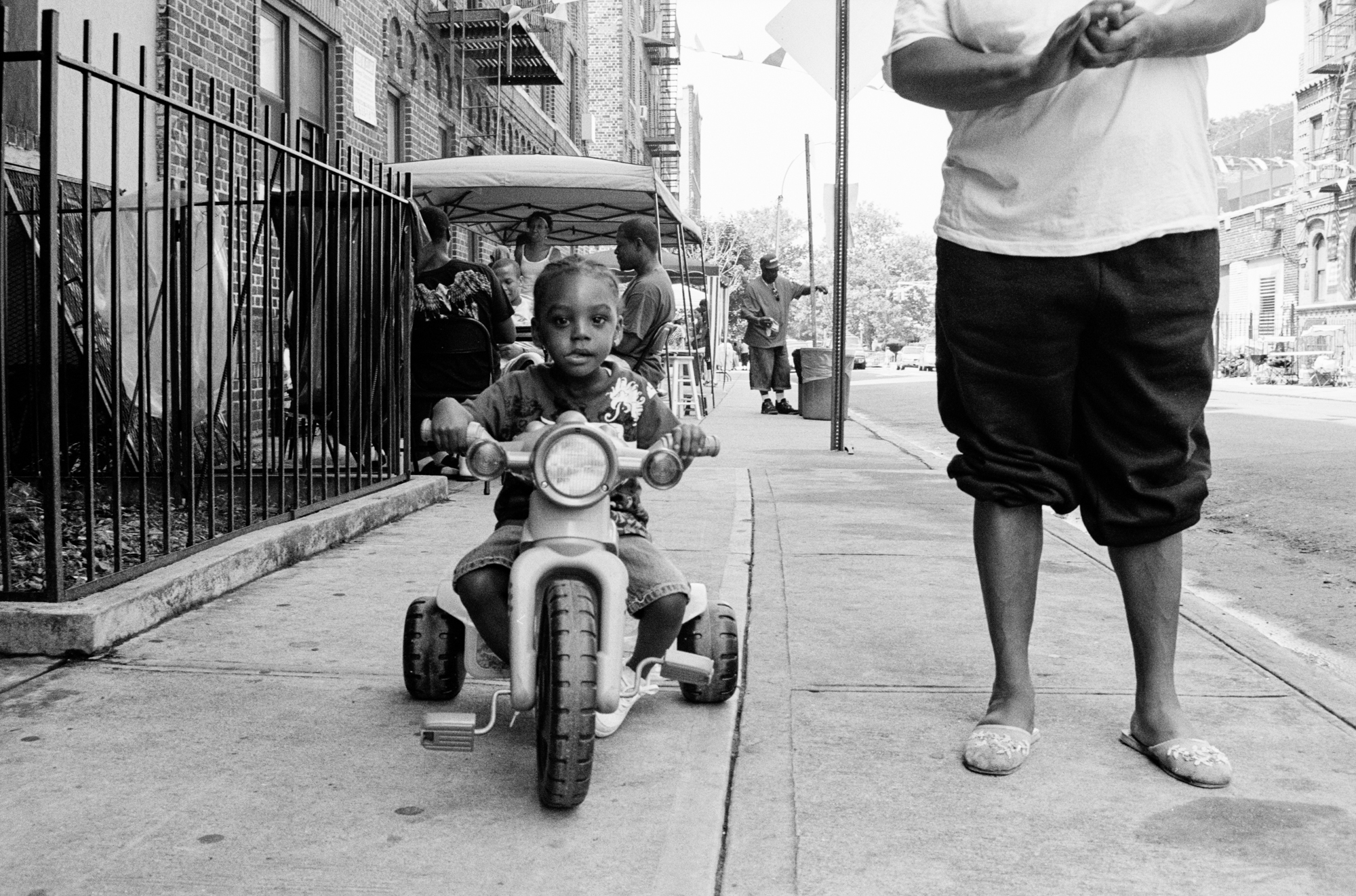 Black and white image of a young child riding a motorcycle on a city street, surrounded by pedestrians and buildings.