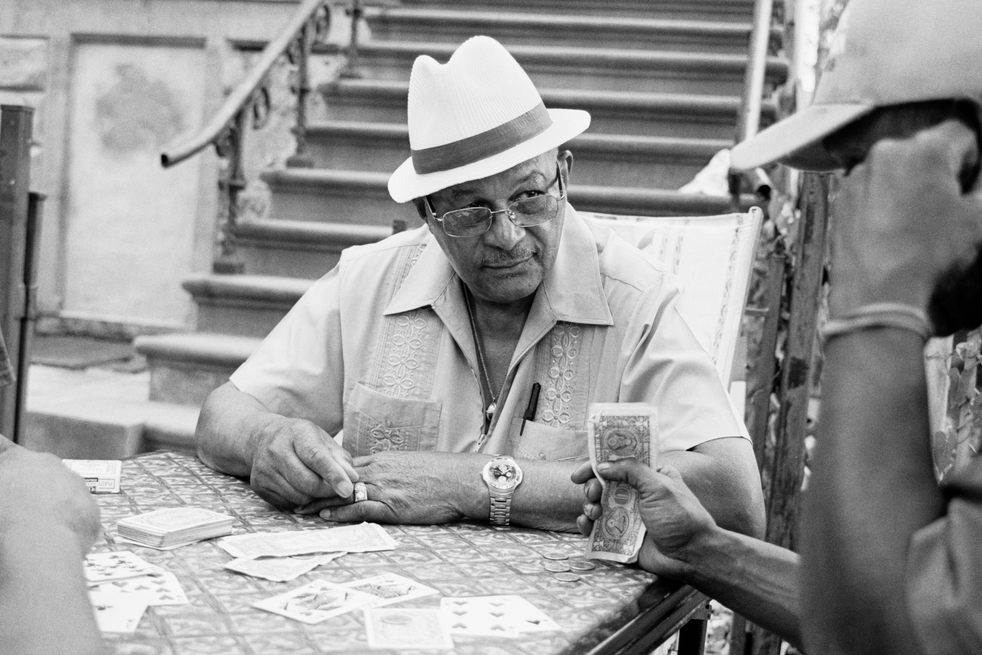 A middle-aged man wearing a white hat, glasses, and a light-coloured shirt sits at a table, intently studying papers and documents.