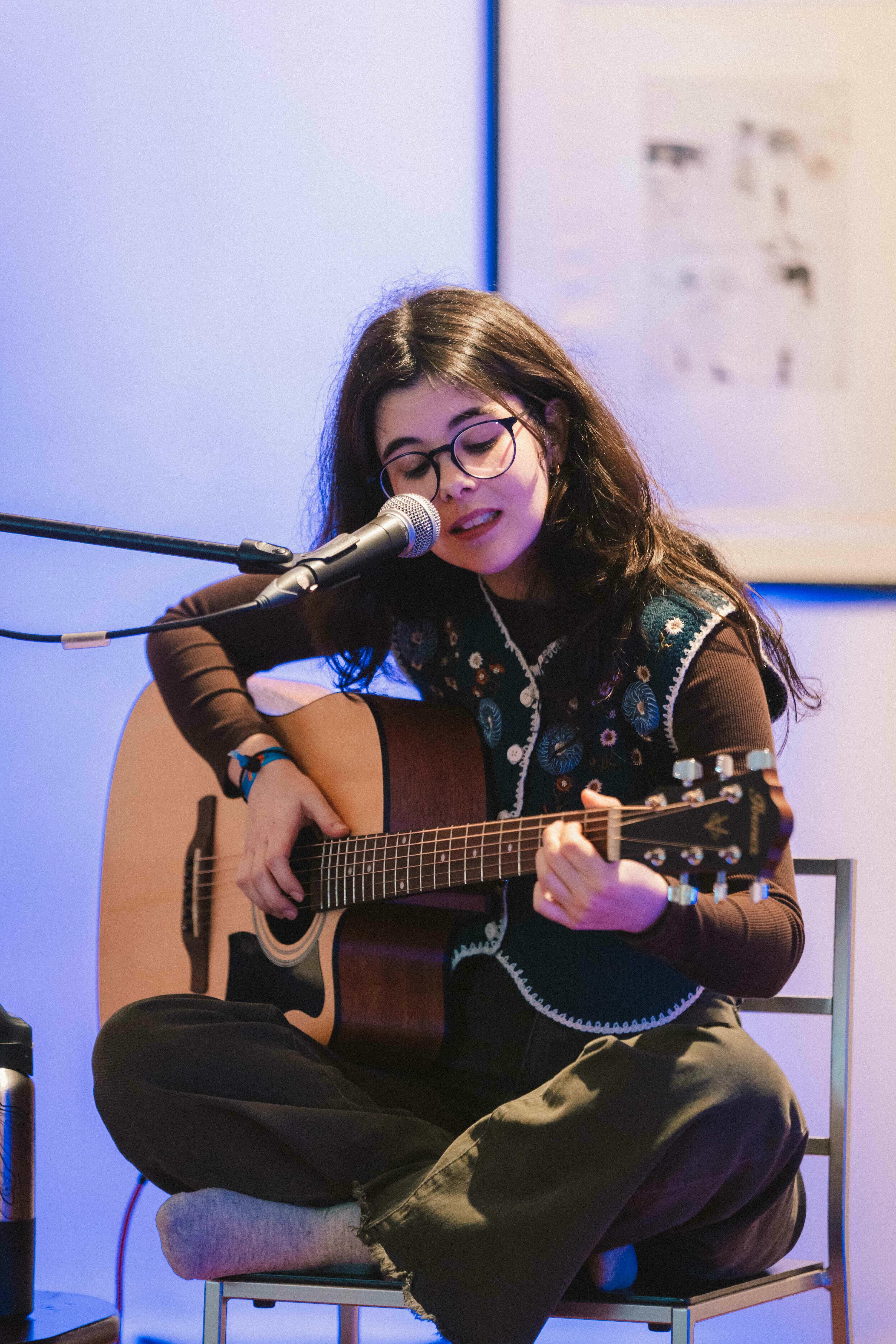 Young woman with long brown hair wearing glasses and a floral shirt, sitting on a chair and playing an acoustic guitar while singing into a microphone.