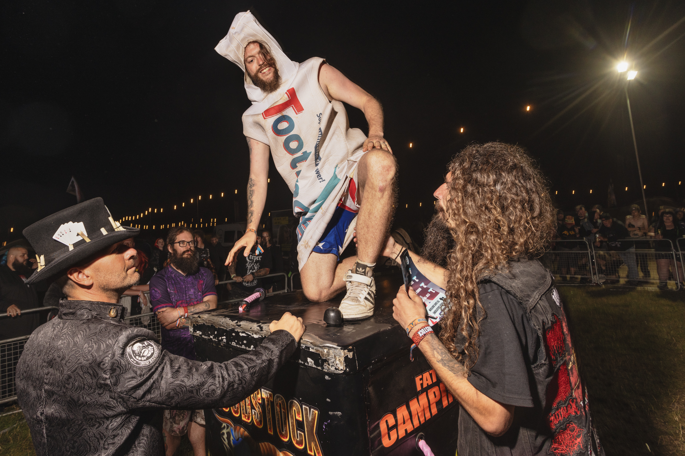 Person in white towel costume with "TOOL" text being lifted by crowd at night-time outdoor event with police officer present.