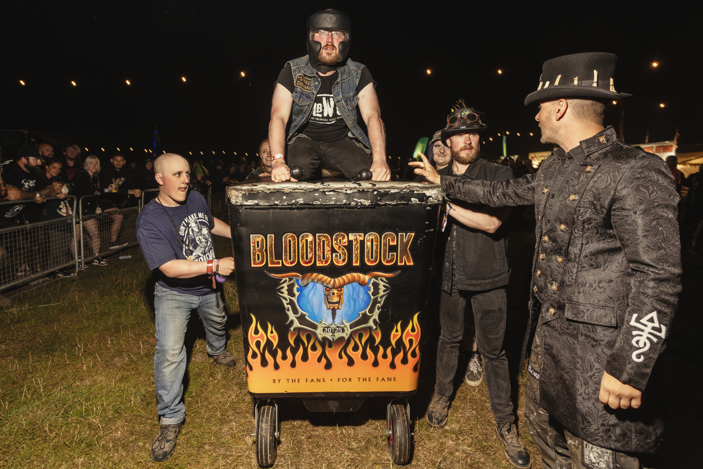 Man in hat standing behind "Bloodstock" barbecue grill with flame graphics, surrounded by three people in dark clothing at nighttime event.