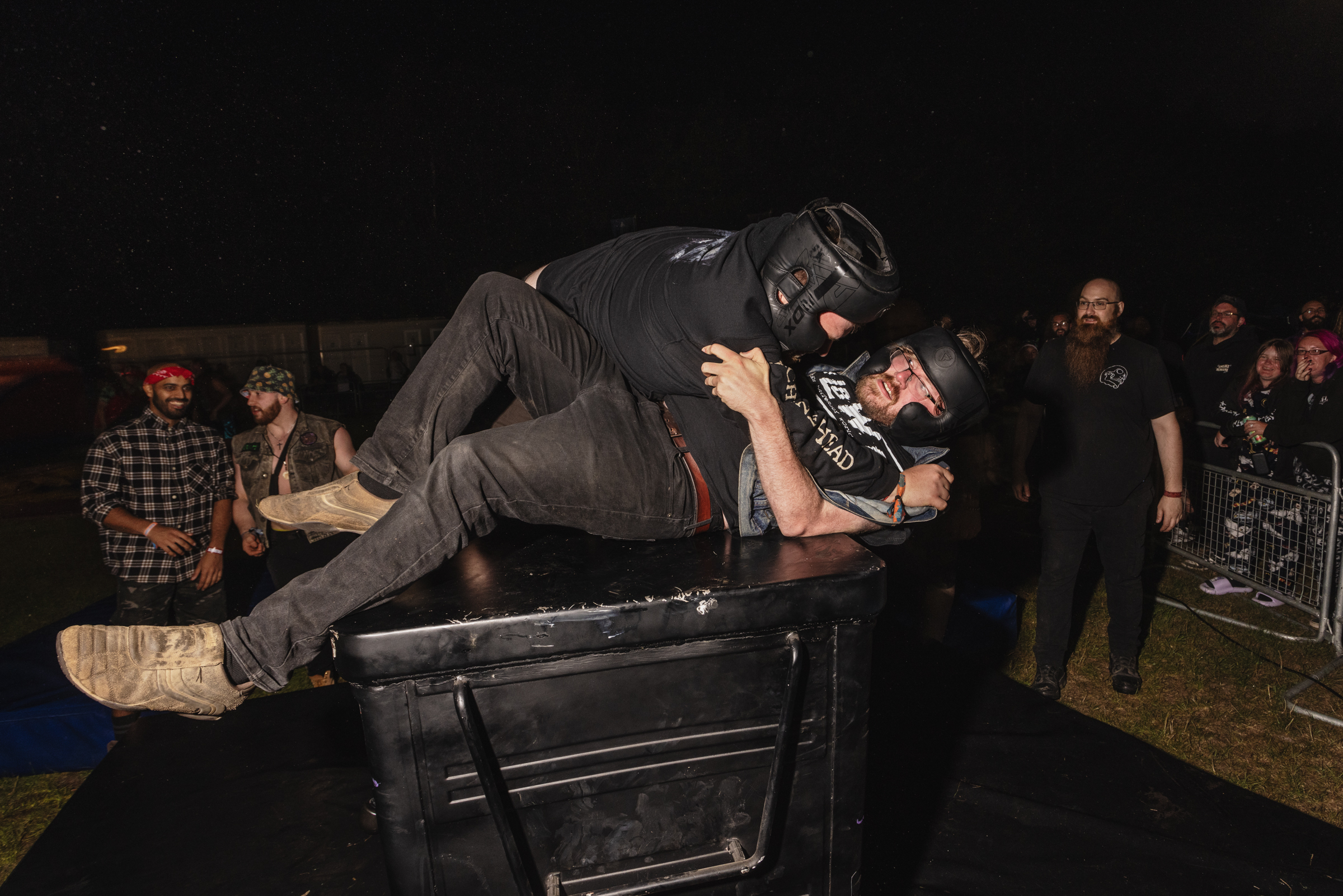 Wrestler in dark clothing performs move on opponent lying on black table, surrounded by crowd in dimly lit venue.