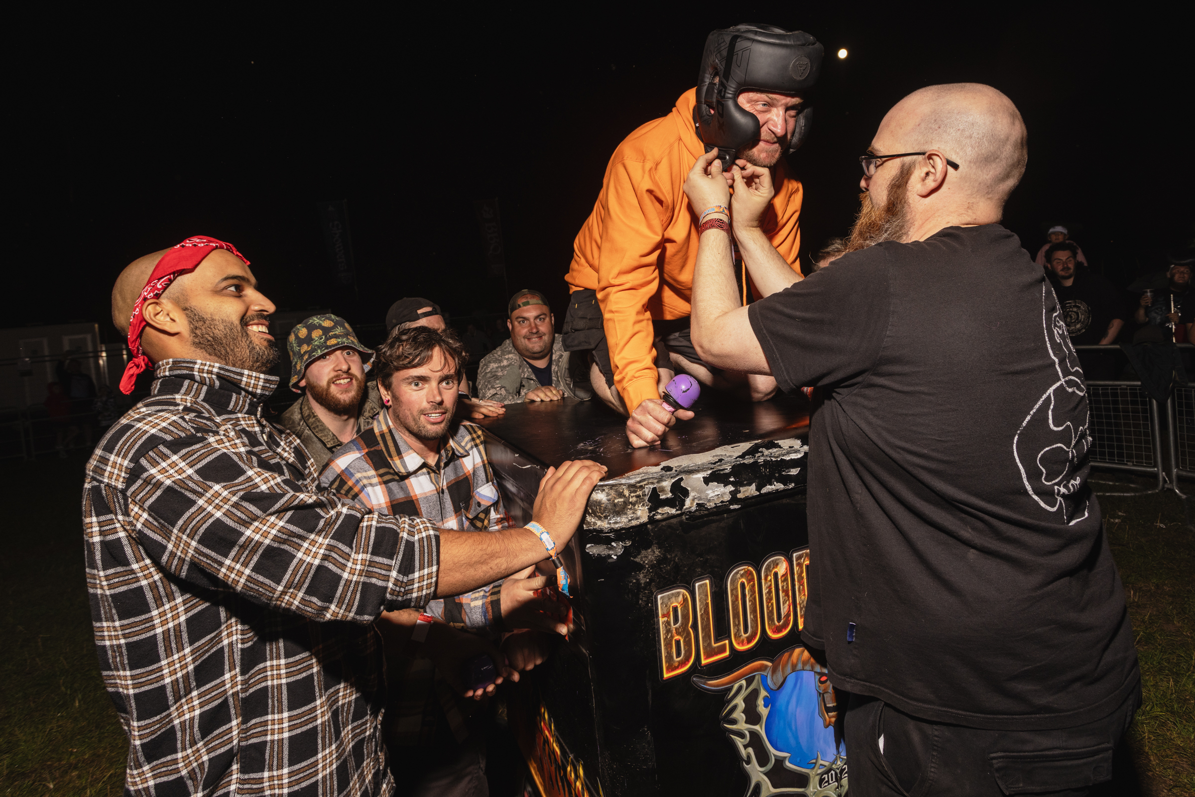 Group of men gathered around table at night, one in orange jumpsuit being restrained, others in casual clothing watching.