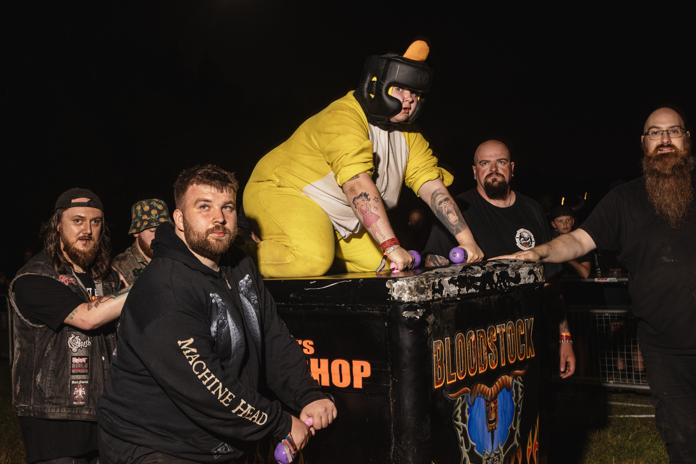 Man in bright yellow tracksuit and beanie leaning over table whilst four bearded men in dark clothing watch from behind.