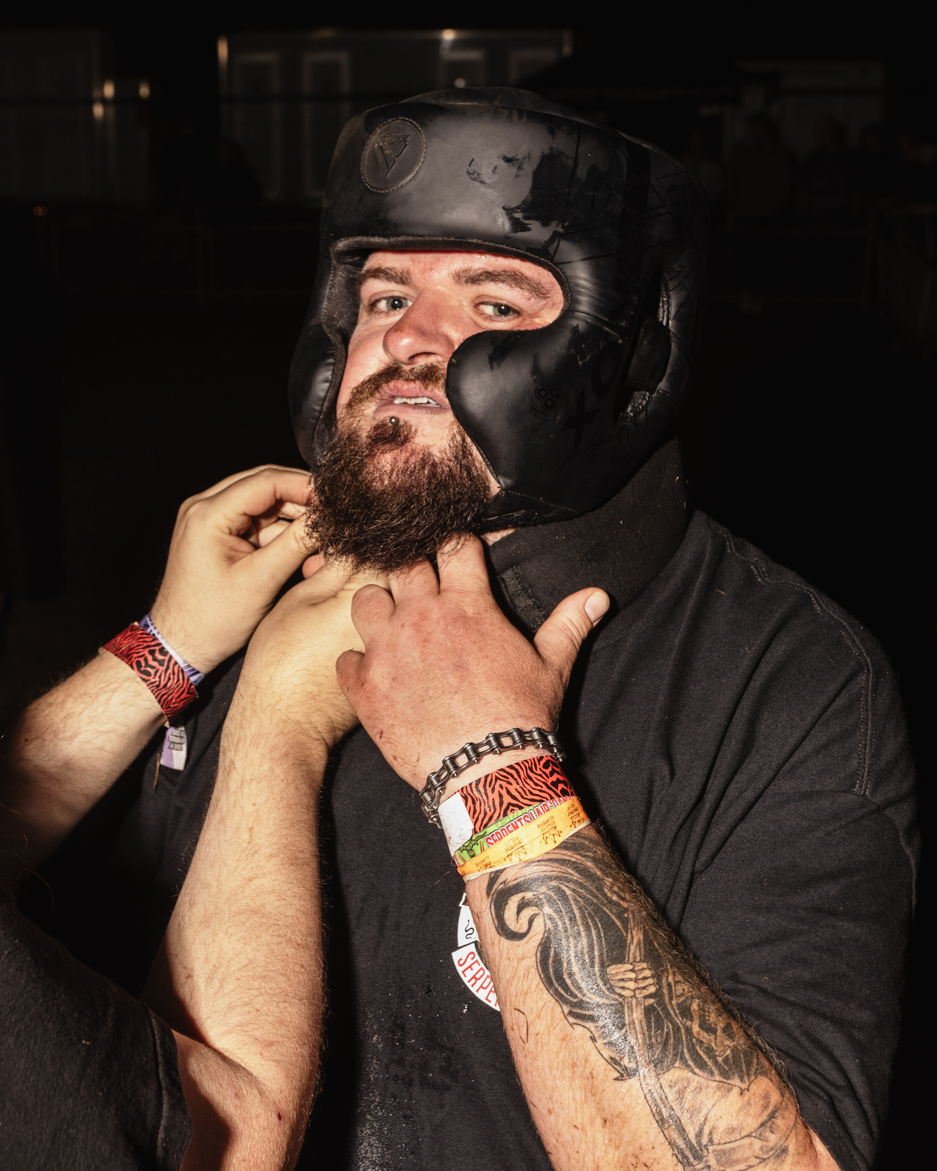 Bearded man removing black helmet, wearing dark clothing with tattooed arms and red wristbands in dimly lit setting.