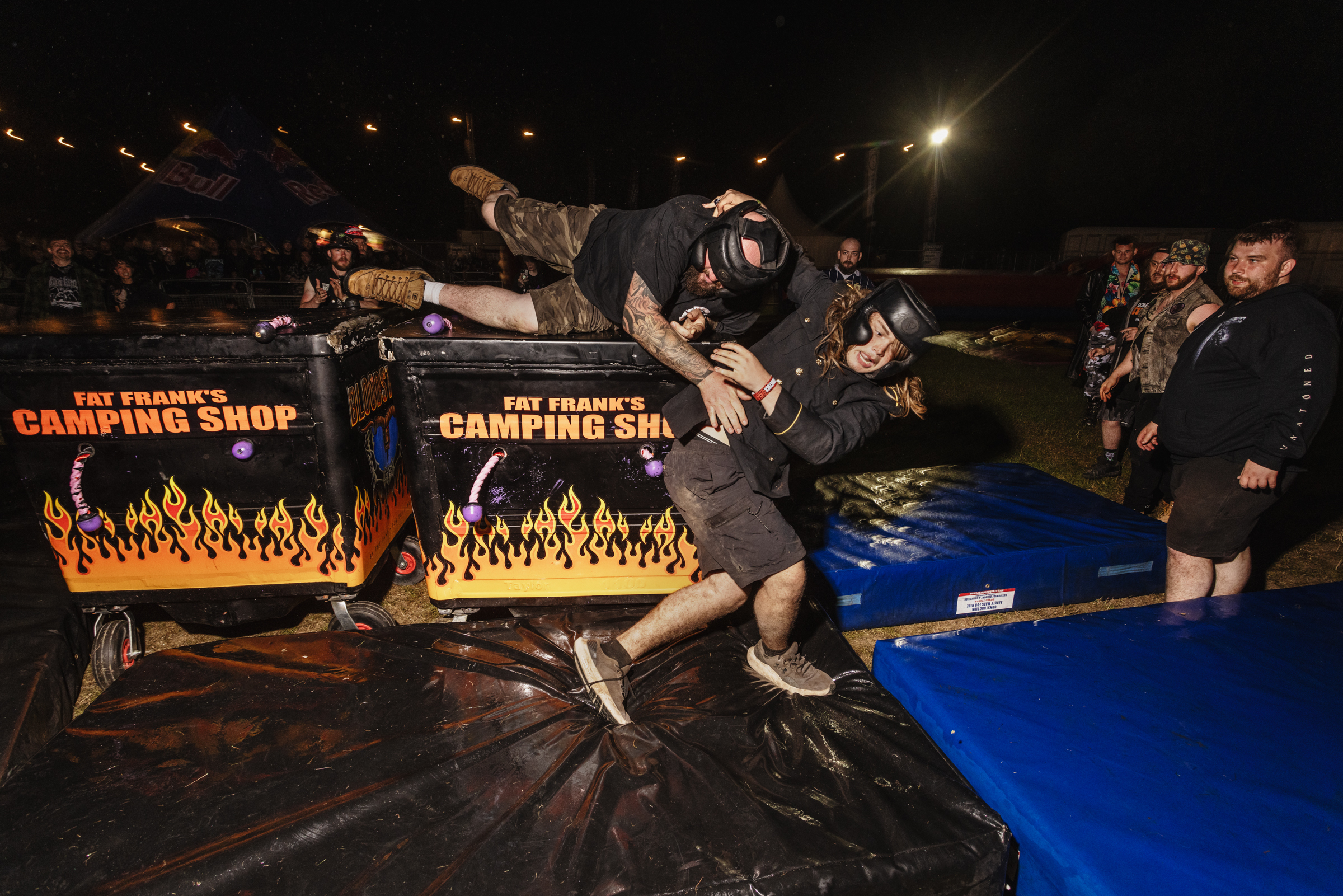 Wrestler in mask and camouflage gear attacking opponent through flaming table at indoor wrestling event with crowd watching.