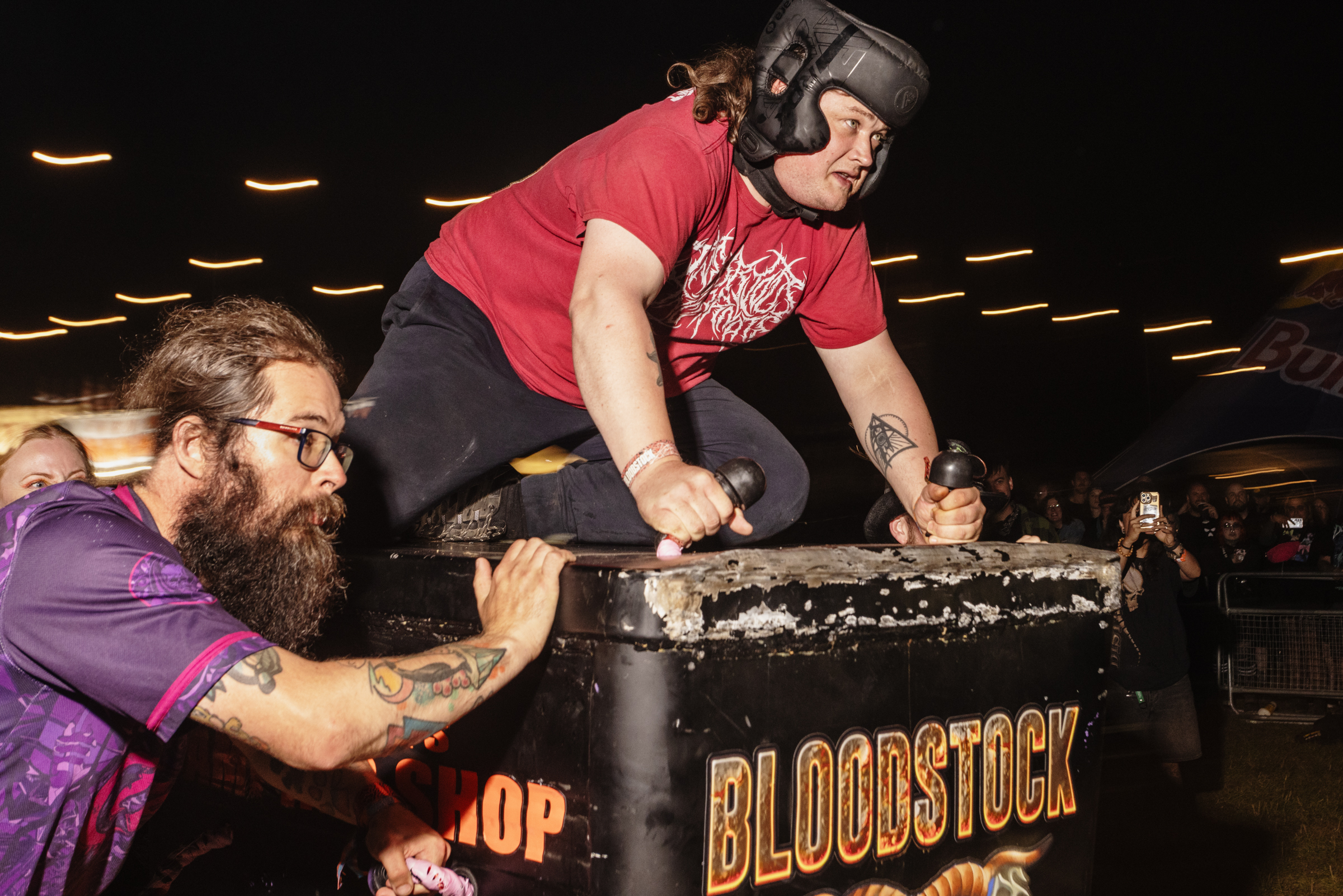 Two wrestlers grappling over wrestling ring ropes. One wears red shirt and black shorts, other in purple with beard and glasses. "BLOODSTOCK" visible on ring apron.