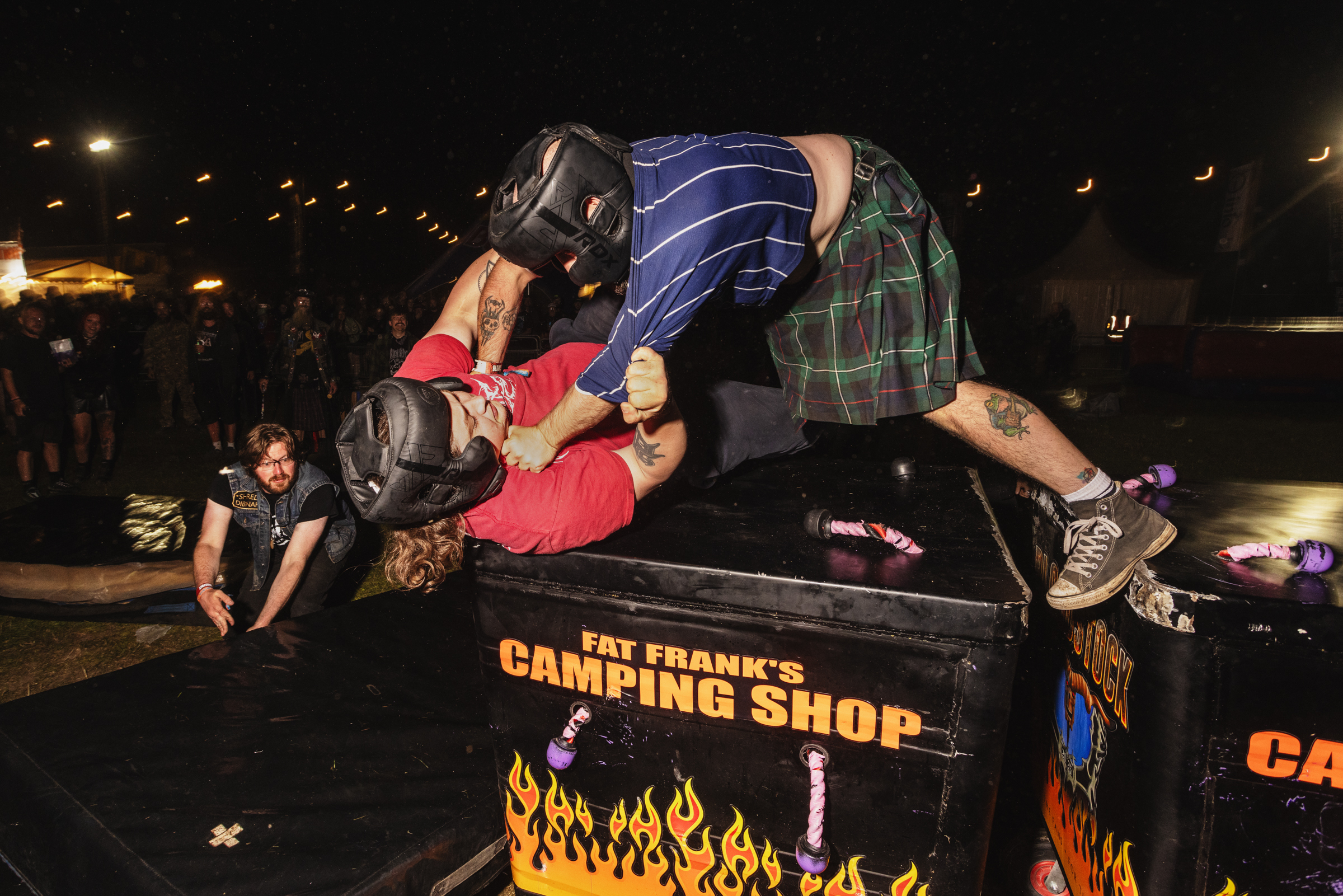 Wrestling match on black table with flame graphics; wrestlers in red and blue shirts grappling whilst crowd watches in background.