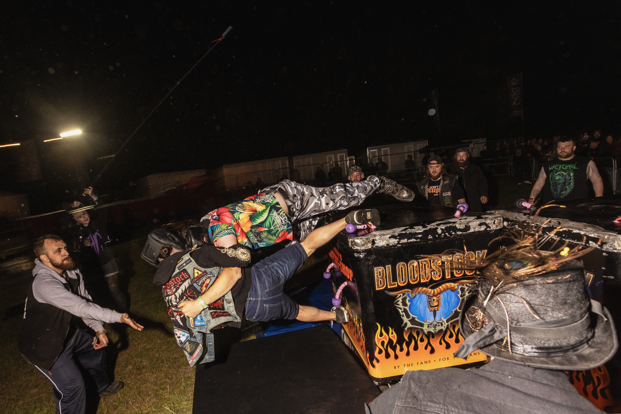 Demolished car covered in stickers and graffiti surrounded by crowd at nighttime event, with bright light illuminating dark scene.