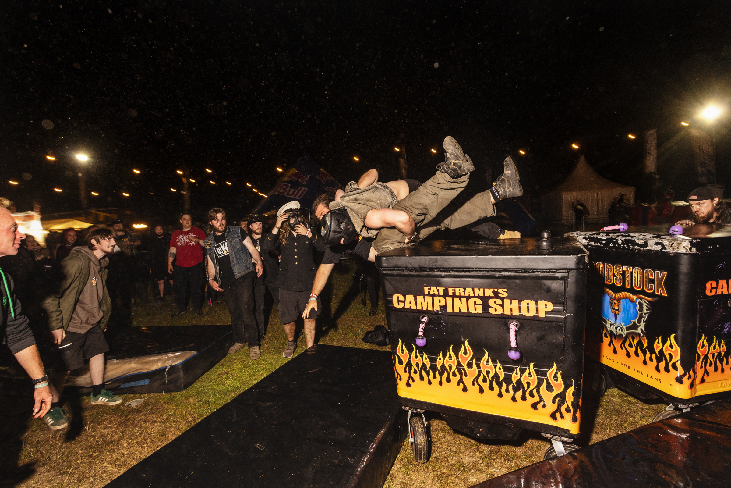 Man leaping horizontally over black and yellow camping shop stall at night outdoor event with crowd watching in background.