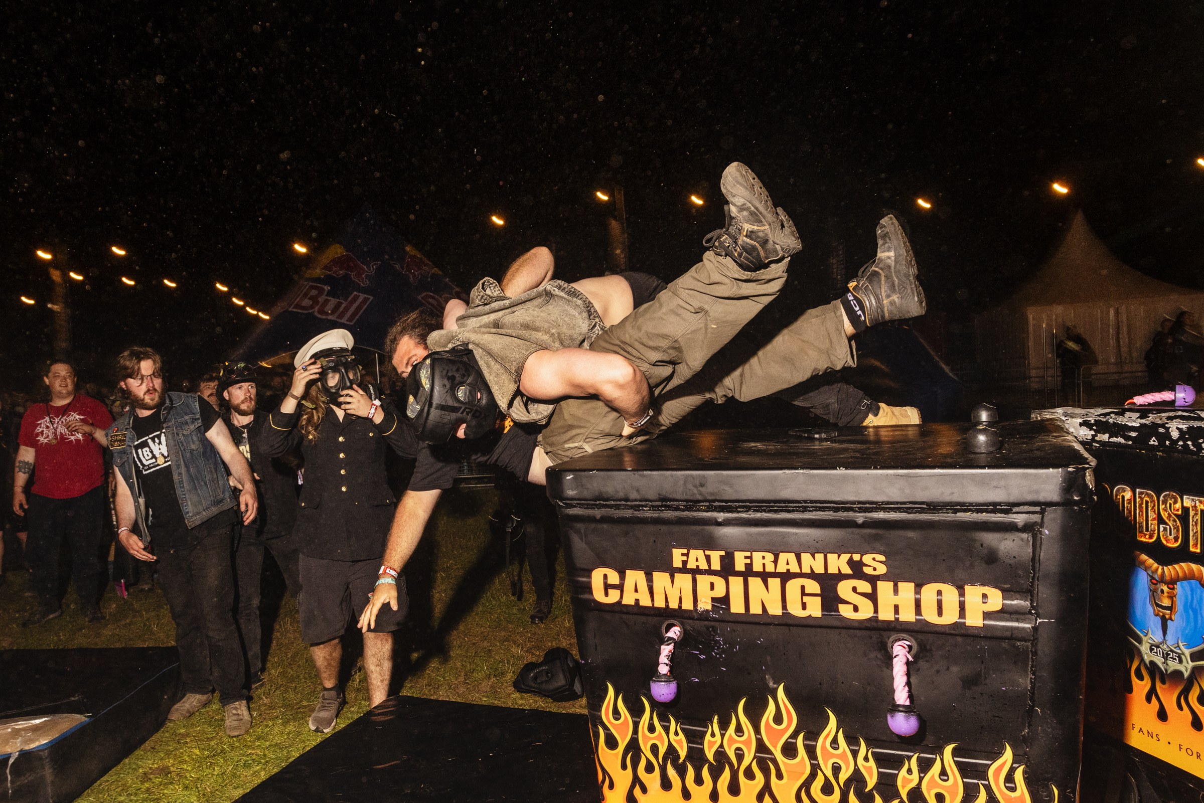Person in camouflage clothing being flipped backwards over black trailer with "Fat Frank's Camping Shop" text and flame graphics at night event.