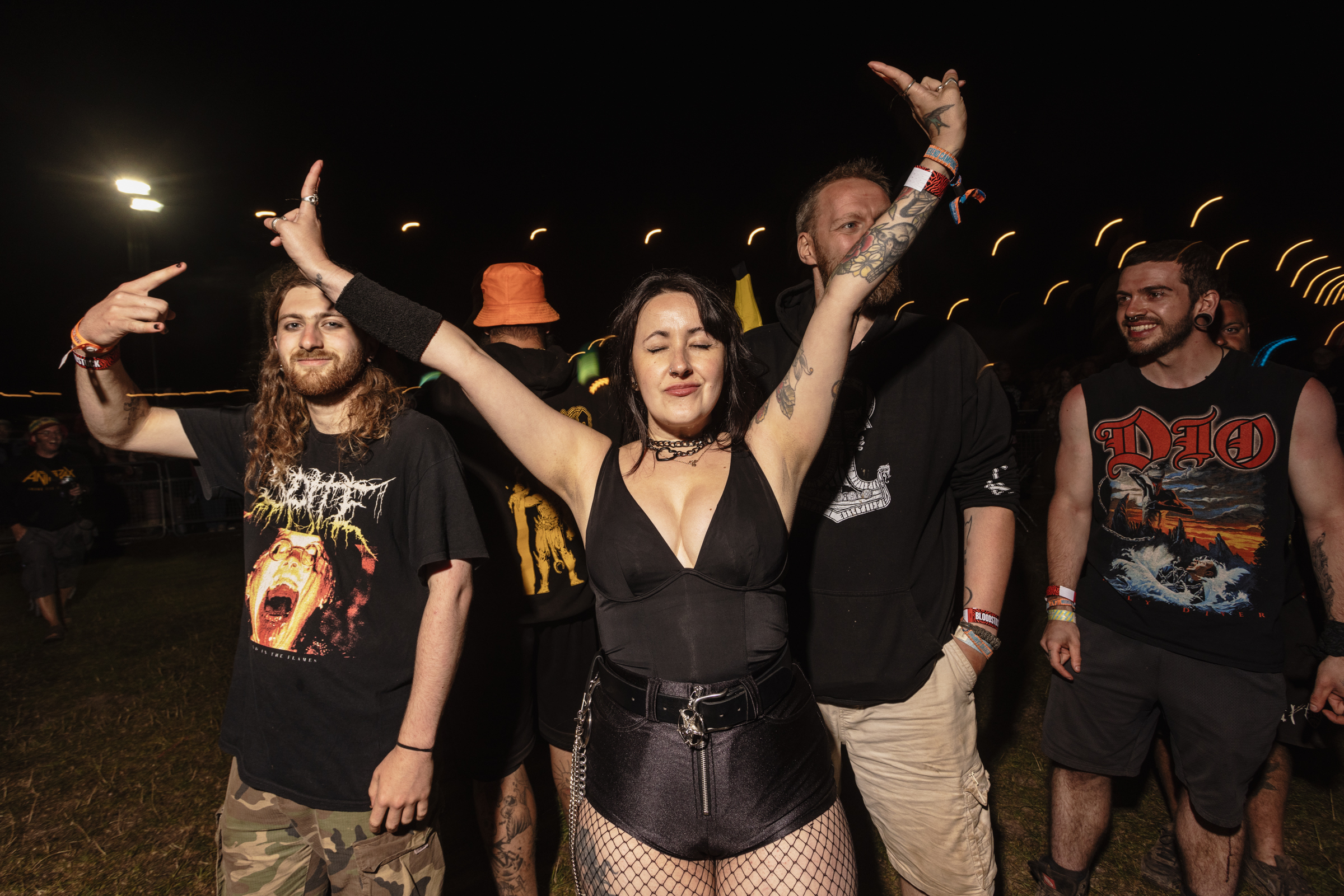 Group of people at outdoor night event, woman in centre wearing black outfit with arms raised, men in band t-shirts around her.