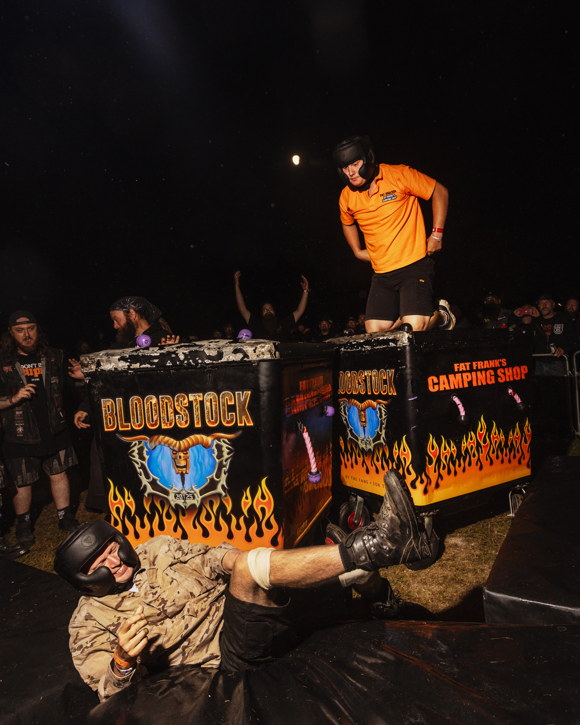 Wrestling performer in orange shirt stands on decorated table with flame designs and "BLOODSTOCK" text whilst another wrestler lies below.