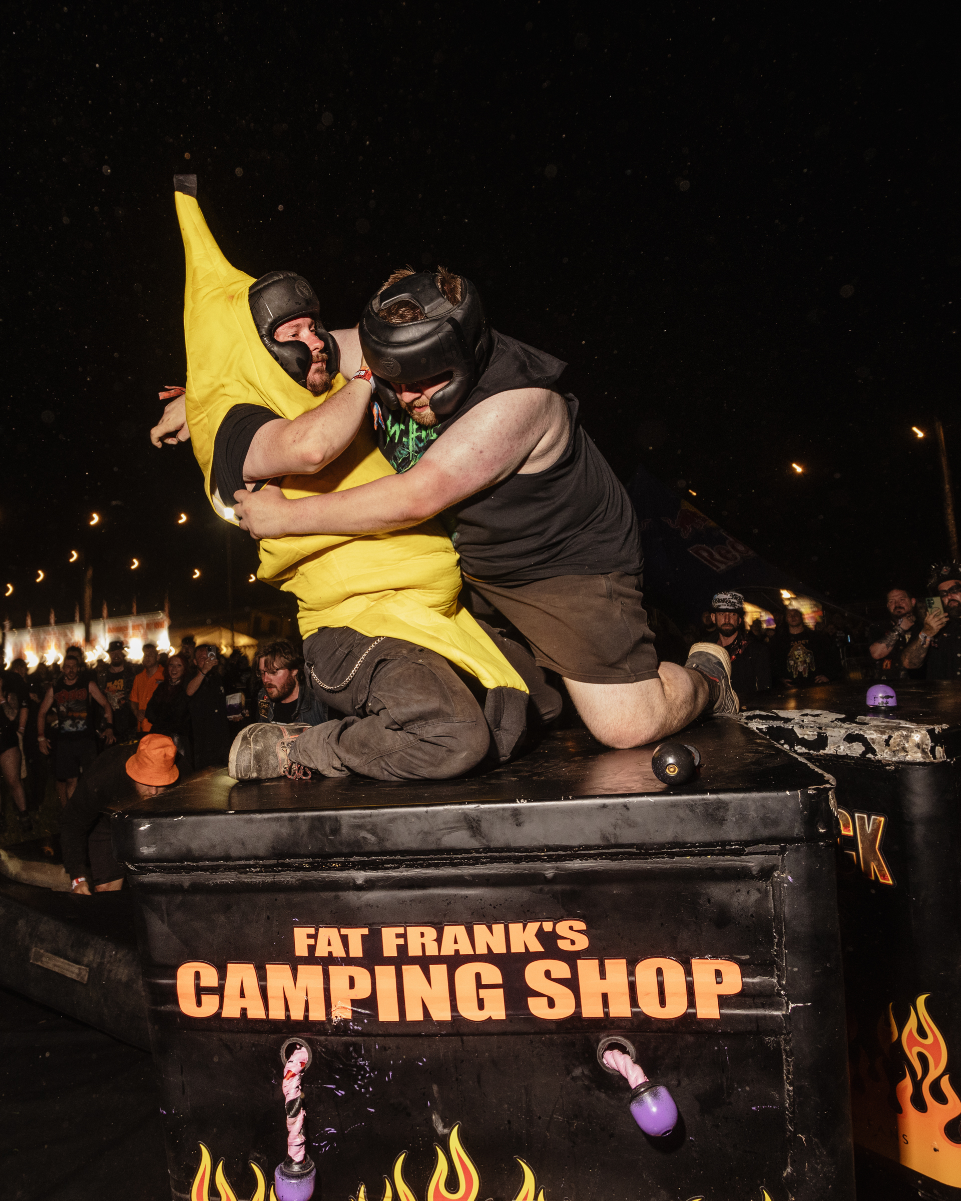 Two wrestlers grappling on stage at night event, one in yellow costume, one in dark clothing, crowd visible in background.