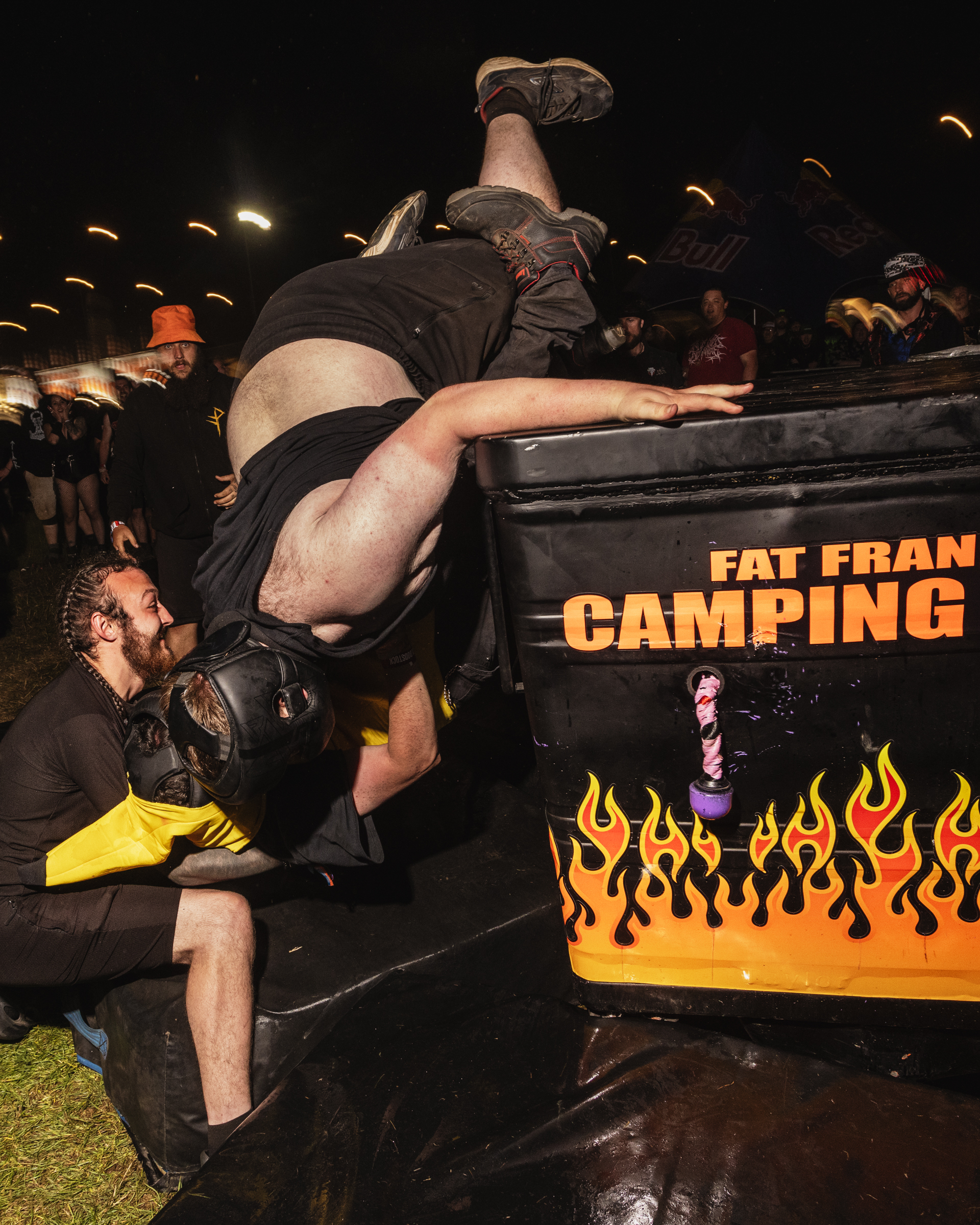Wrestler being thrown backwards over black barrier with orange flame graphics and "Fat Frank Camping" text, crowd watching in background.