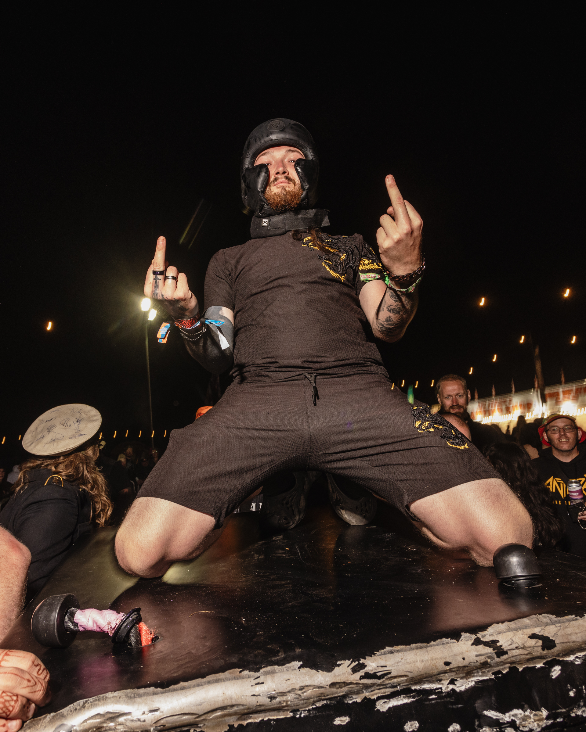 Bearded man in tactical gear and shorts giving middle fingers whilst kneeling on stage at night concert, crowd visible behind.