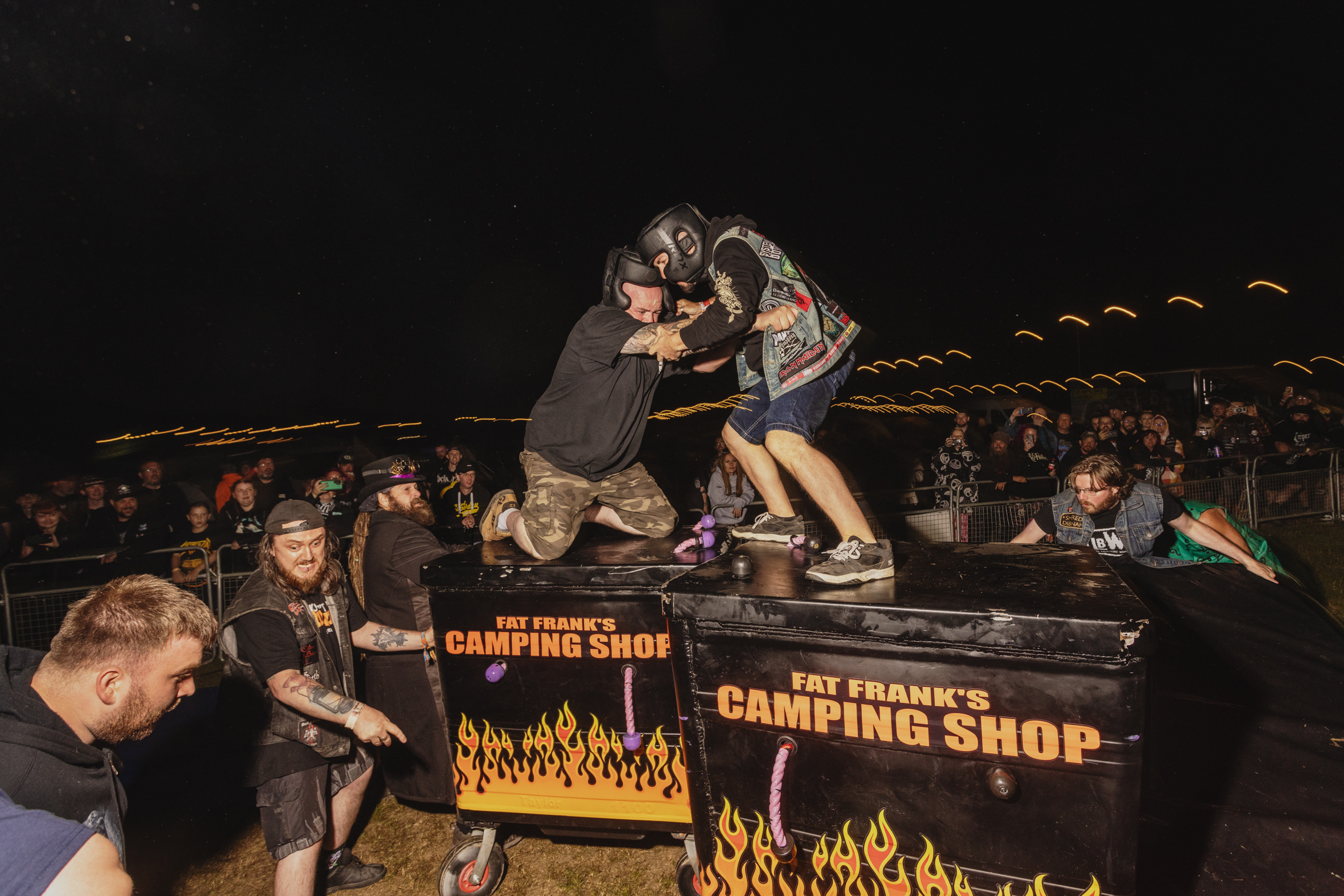 Two wrestlers grappling on black table with flame graphics and "Fat Frank's Camping Shop" text, crowd watching in dark venue.