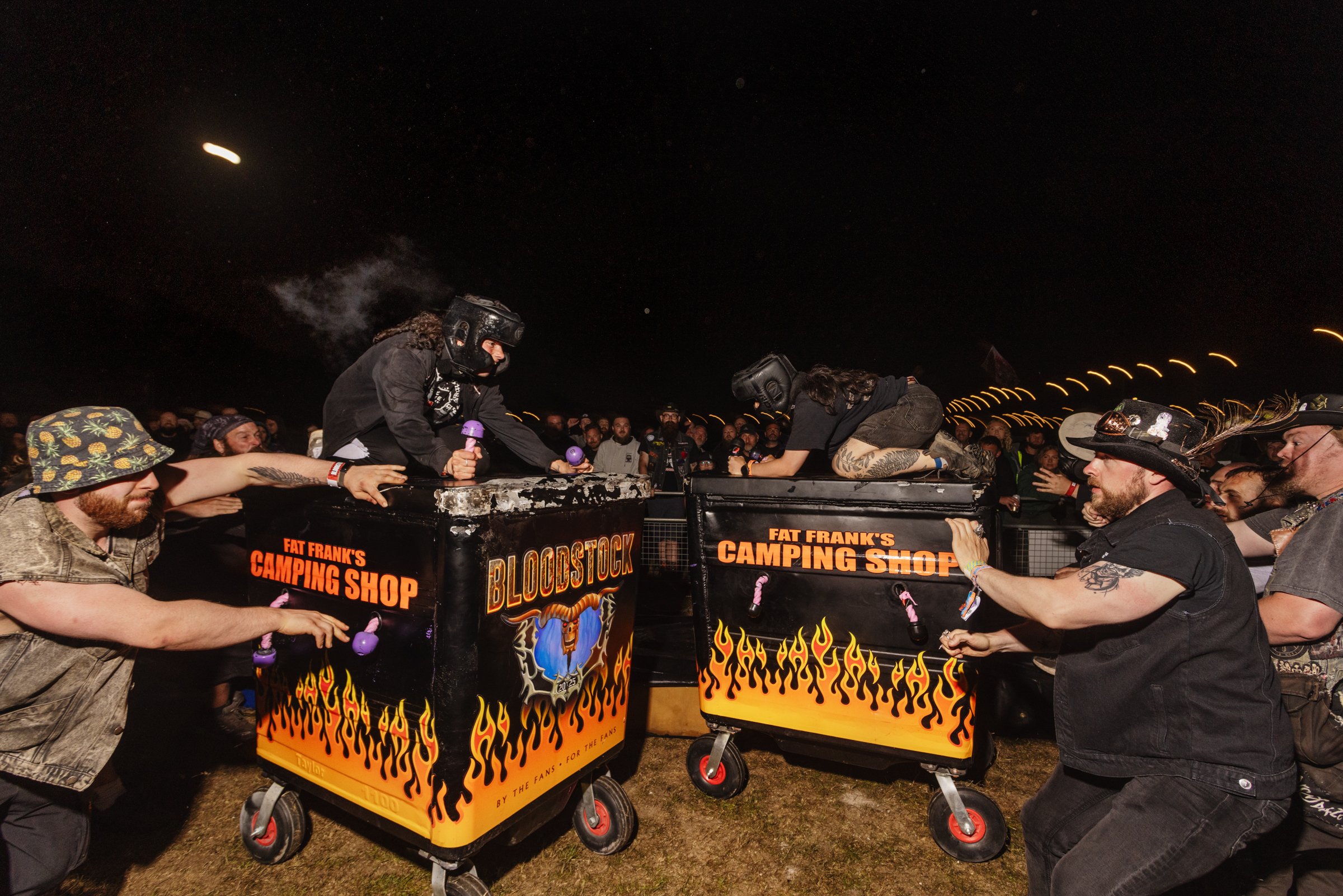 Group of people in camouflage clothing gathered around orange and black decorated camping equipment carts at night outdoors.