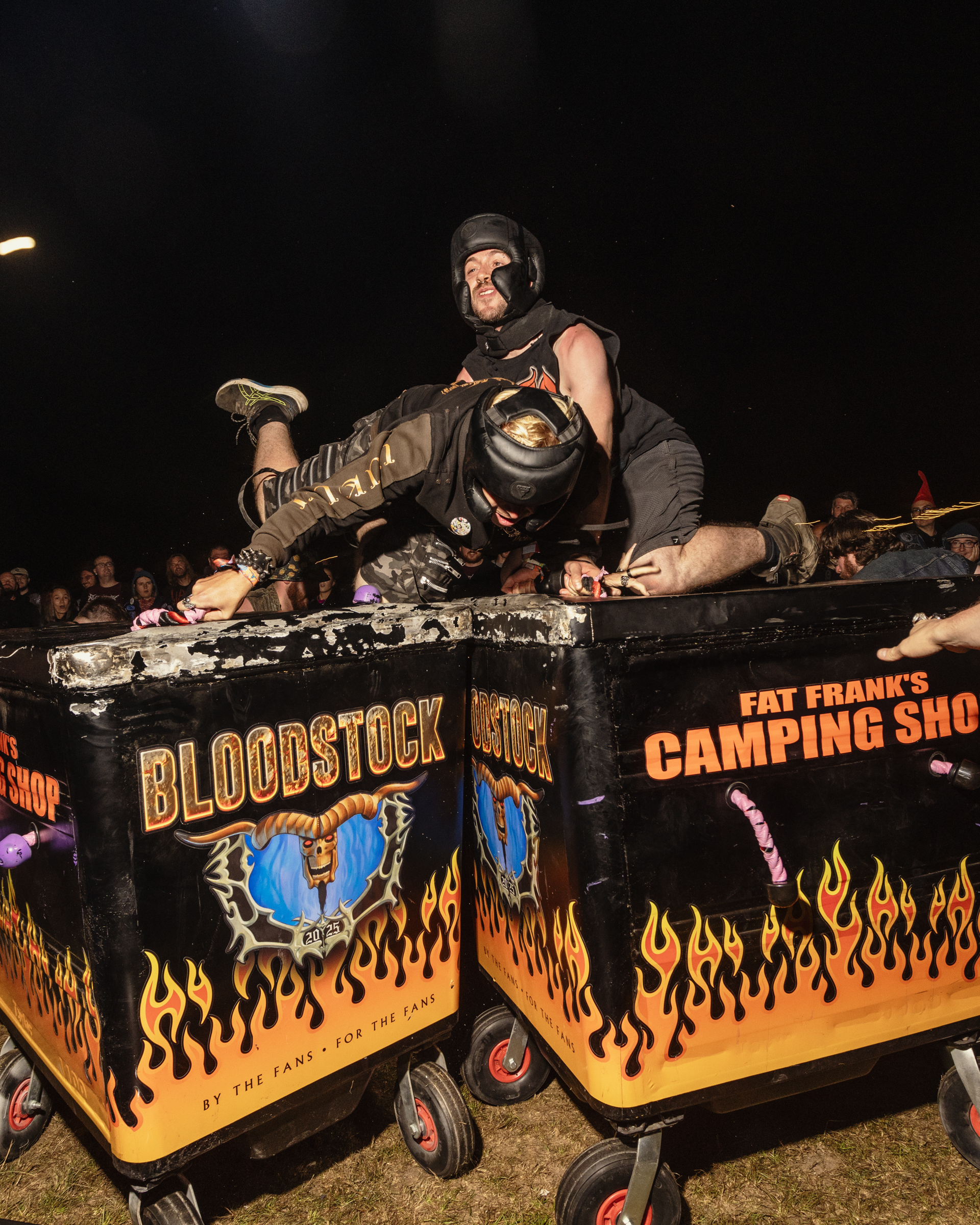 Man in dark clothing sits atop decorated vehicle with orange flame designs and "BLOODSTOCK" text against black night background.