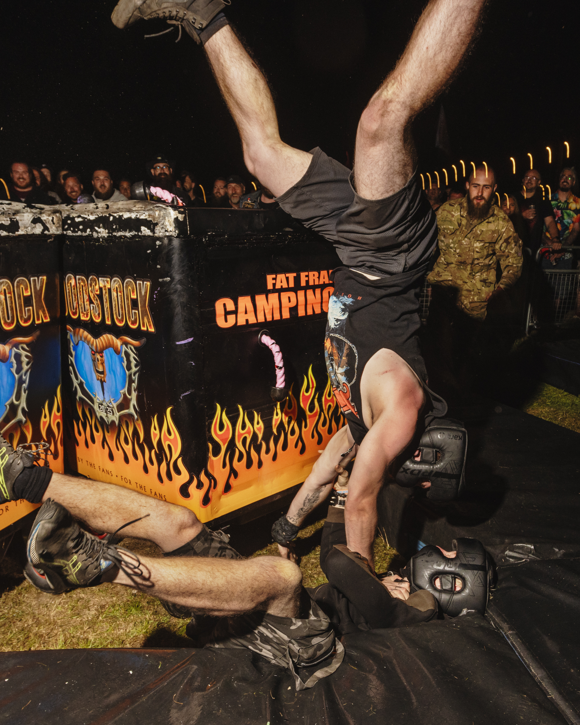 Man performing handstand on black wrestling ring with orange flame graphics, crowd watching in background.