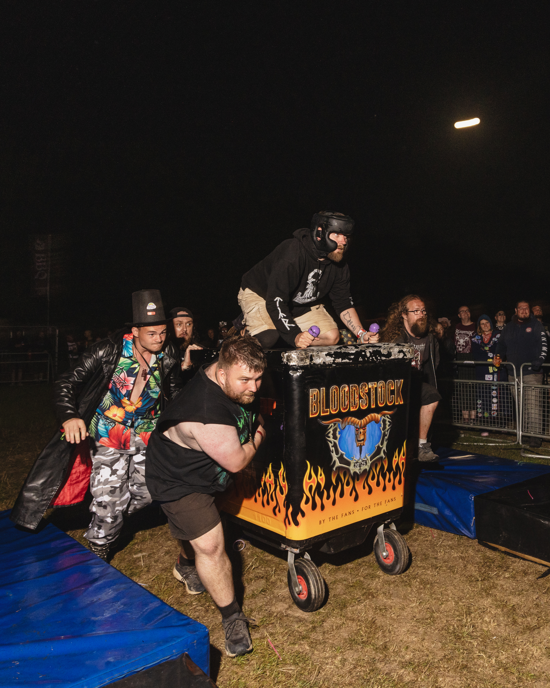 Wrestlers performing at night event with decorated trolley featuring flames and "Bloodstock" text, crowd watching behind barriers.