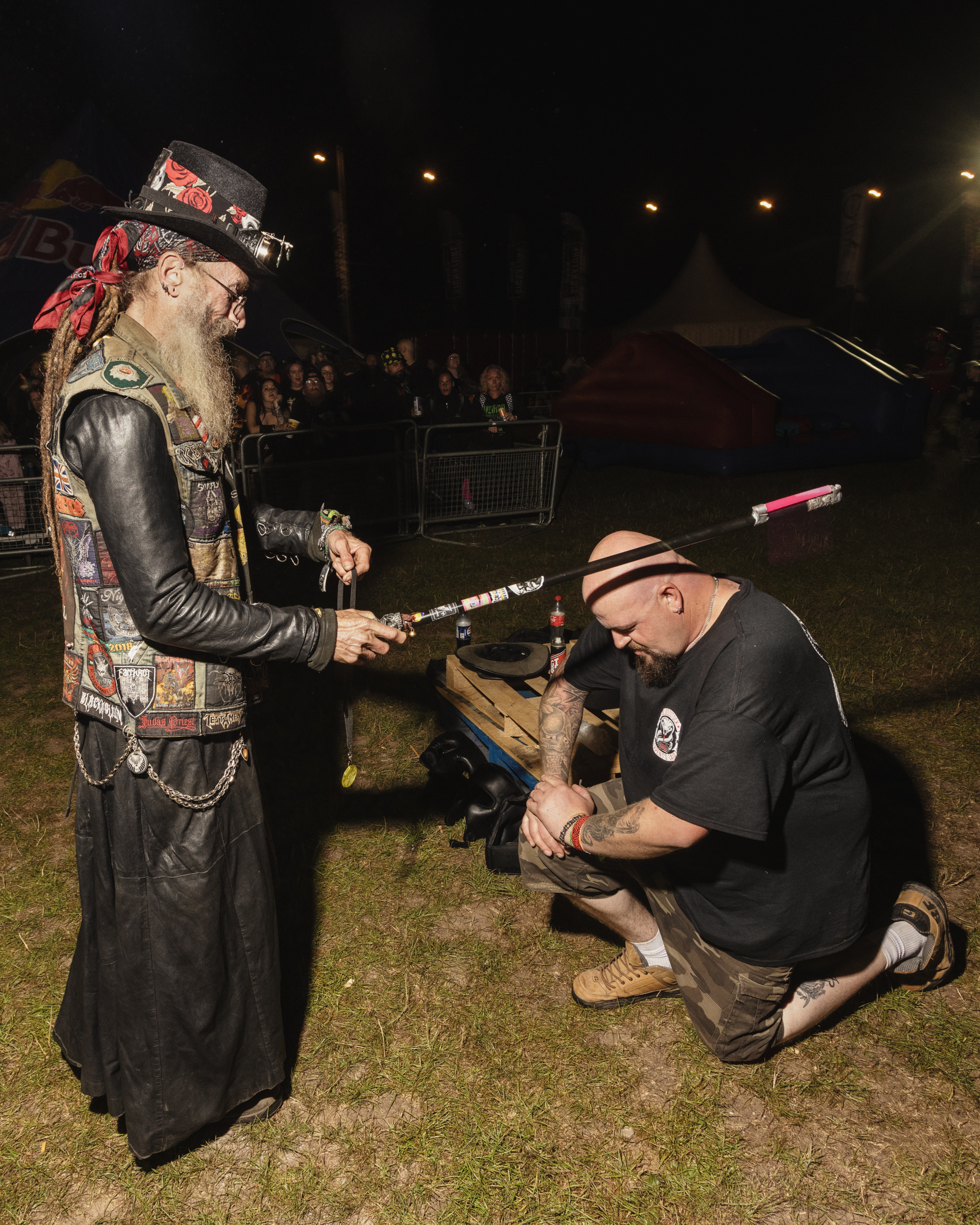 Man in decorated black leather outfit with hat stands whilst another kneels, holding what appears to be a tool or implement at nighttime event.