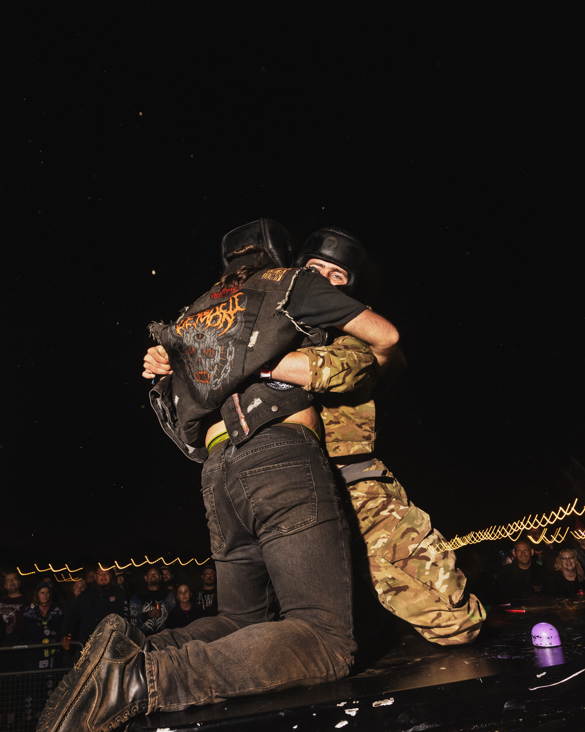 Two performers embracing on stage in dark clothing and face coverings, with audience seated behind chain-link fencing in background.