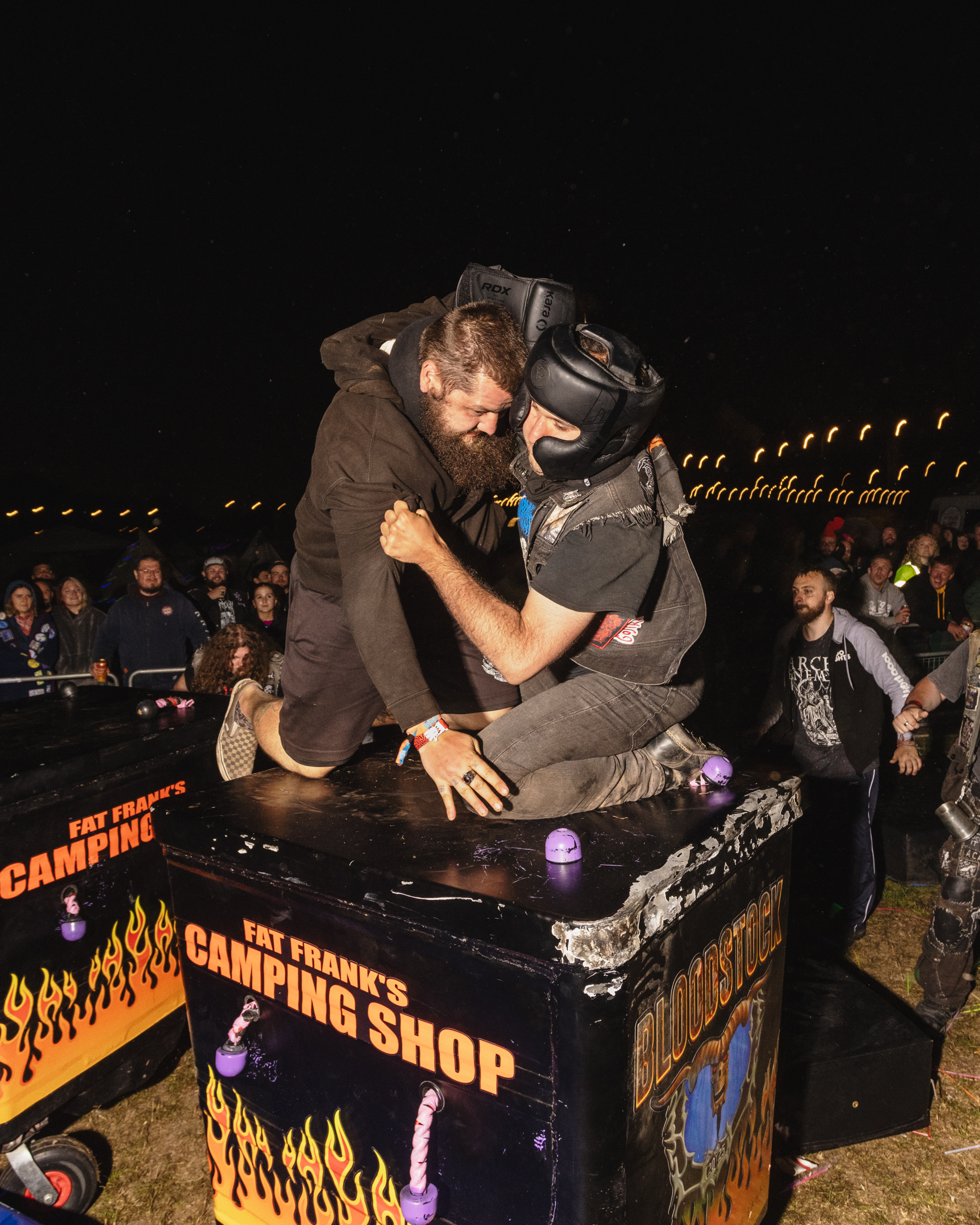 Two wrestlers grappling on a black table with flame graphics and "Campfire Shop" text, surrounded by crowd at night event.