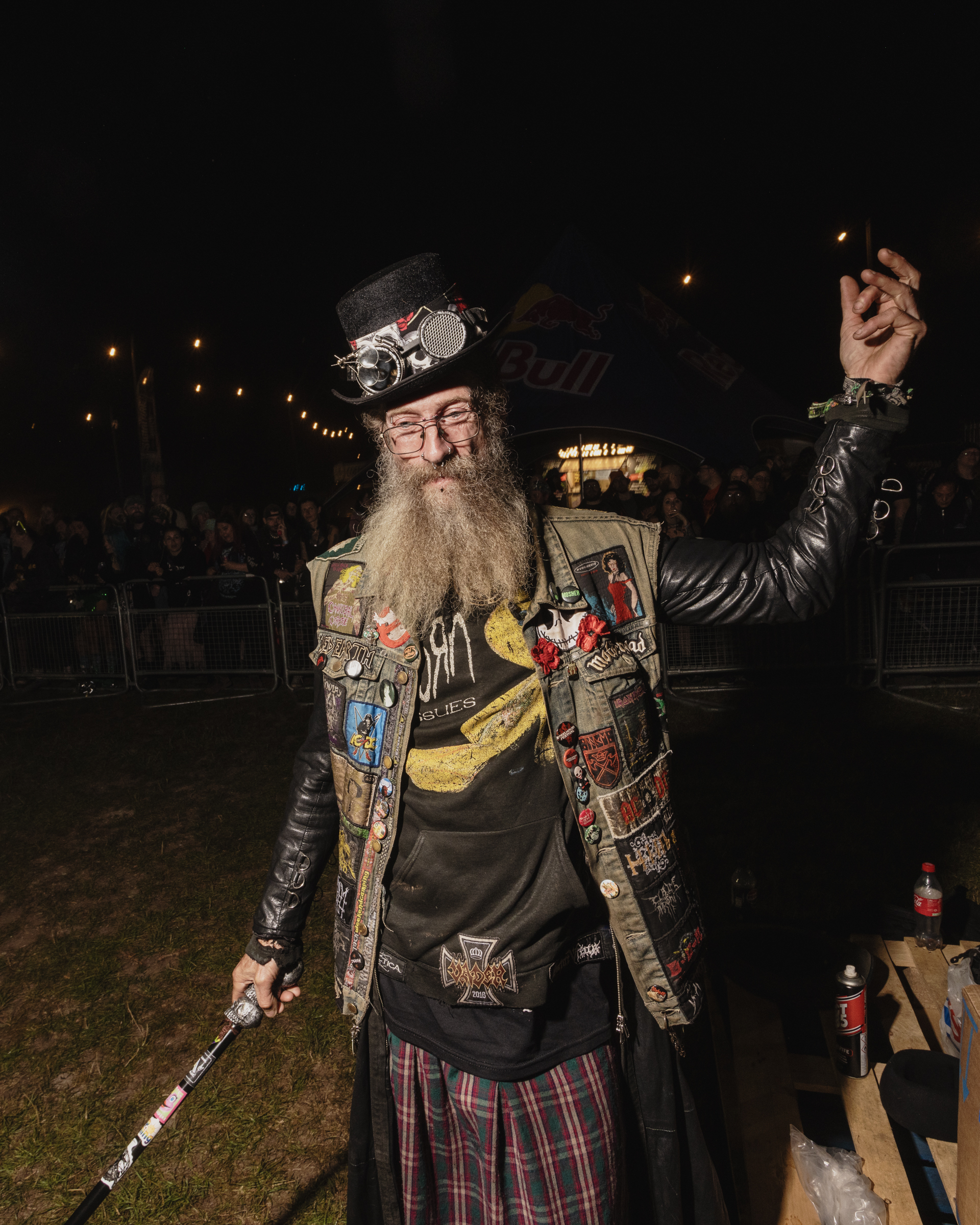 Man with long grey beard wearing decorated steampunk-style jacket with patches, goggles on top hat, raising one arm at night event.