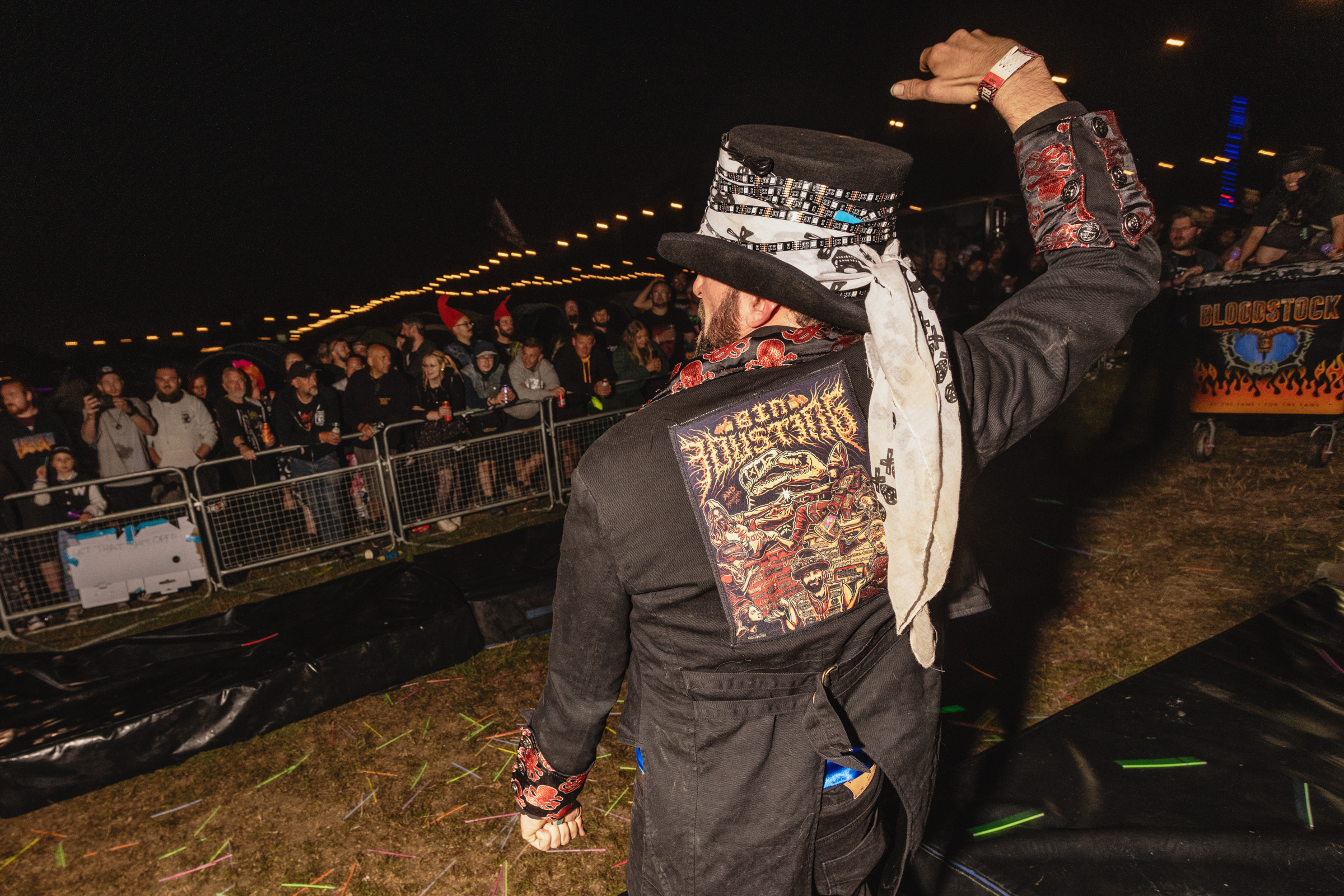 Person in dark clothing with patterned back design performing at night event, crowd watching behind barriers, colourful lights visible.