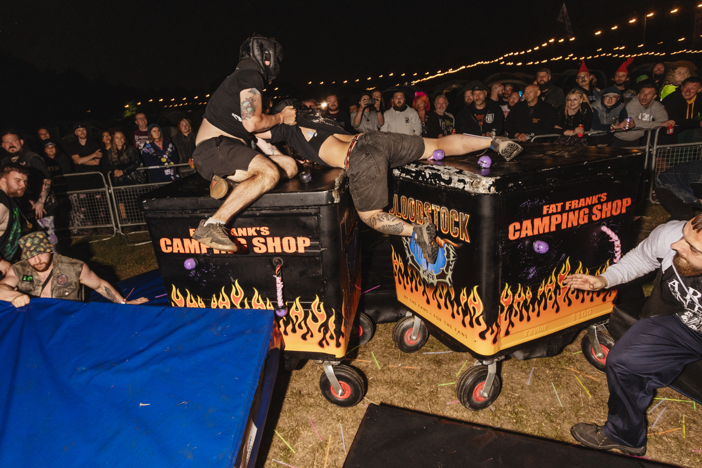 Wrestling match with performer diving from flaming table setup onto opponent, crowd watching from tiered seating under lights.