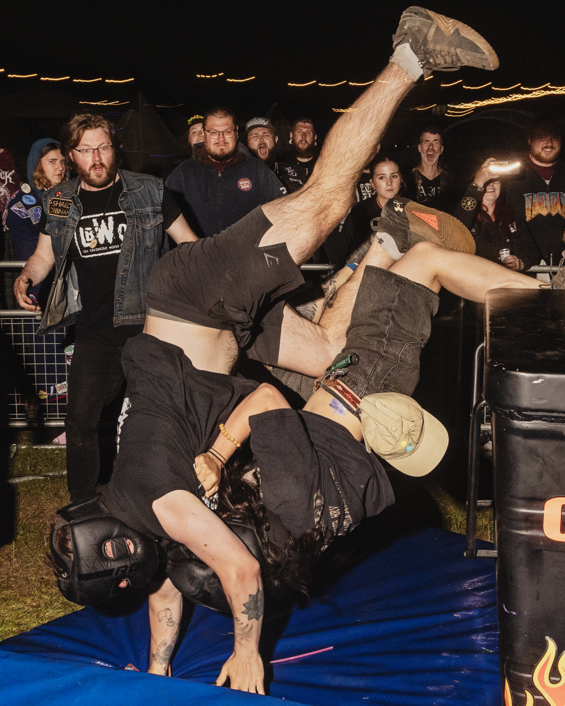 Two wrestlers grappling on blue mat, one upside down with legs raised. Crowd watches from behind barriers in dimly lit venue.