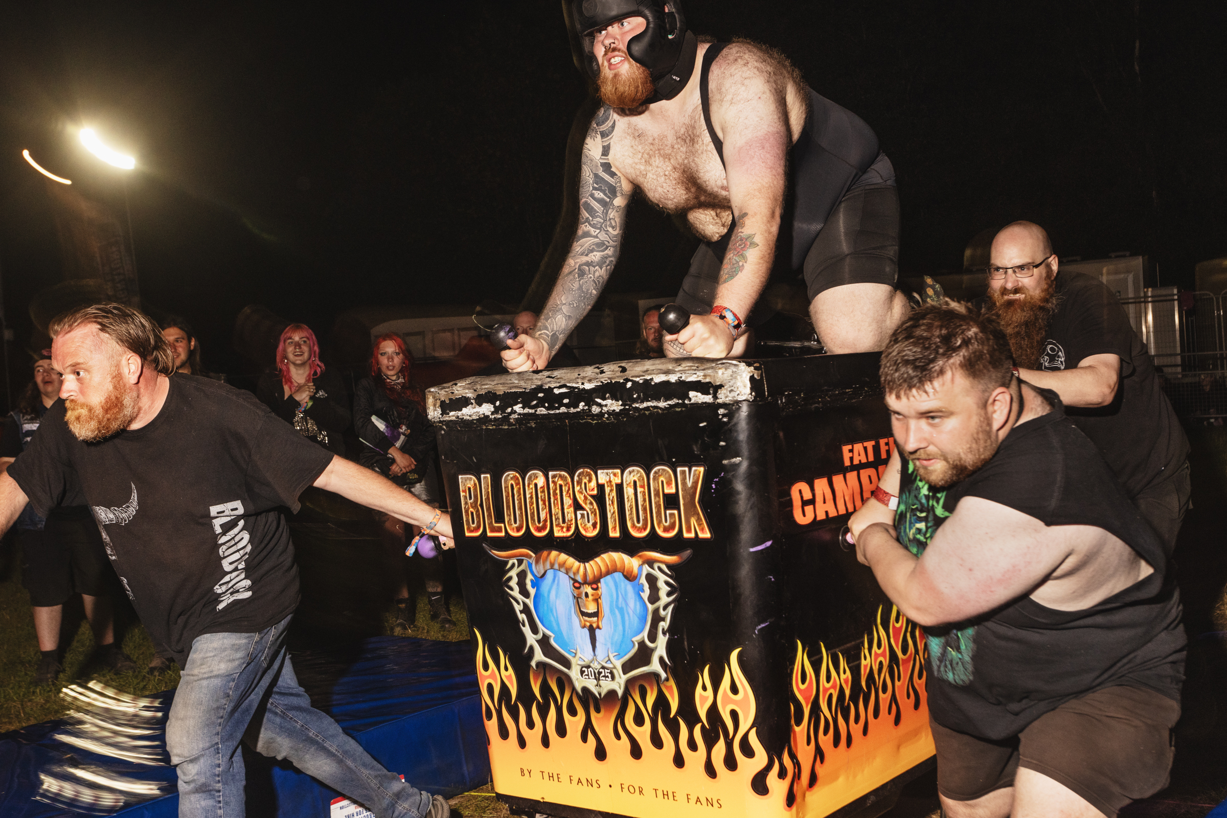 Shirtless wrestler stands on table with "Bloodstock" logo and flame design whilst crowd watches in dark venue