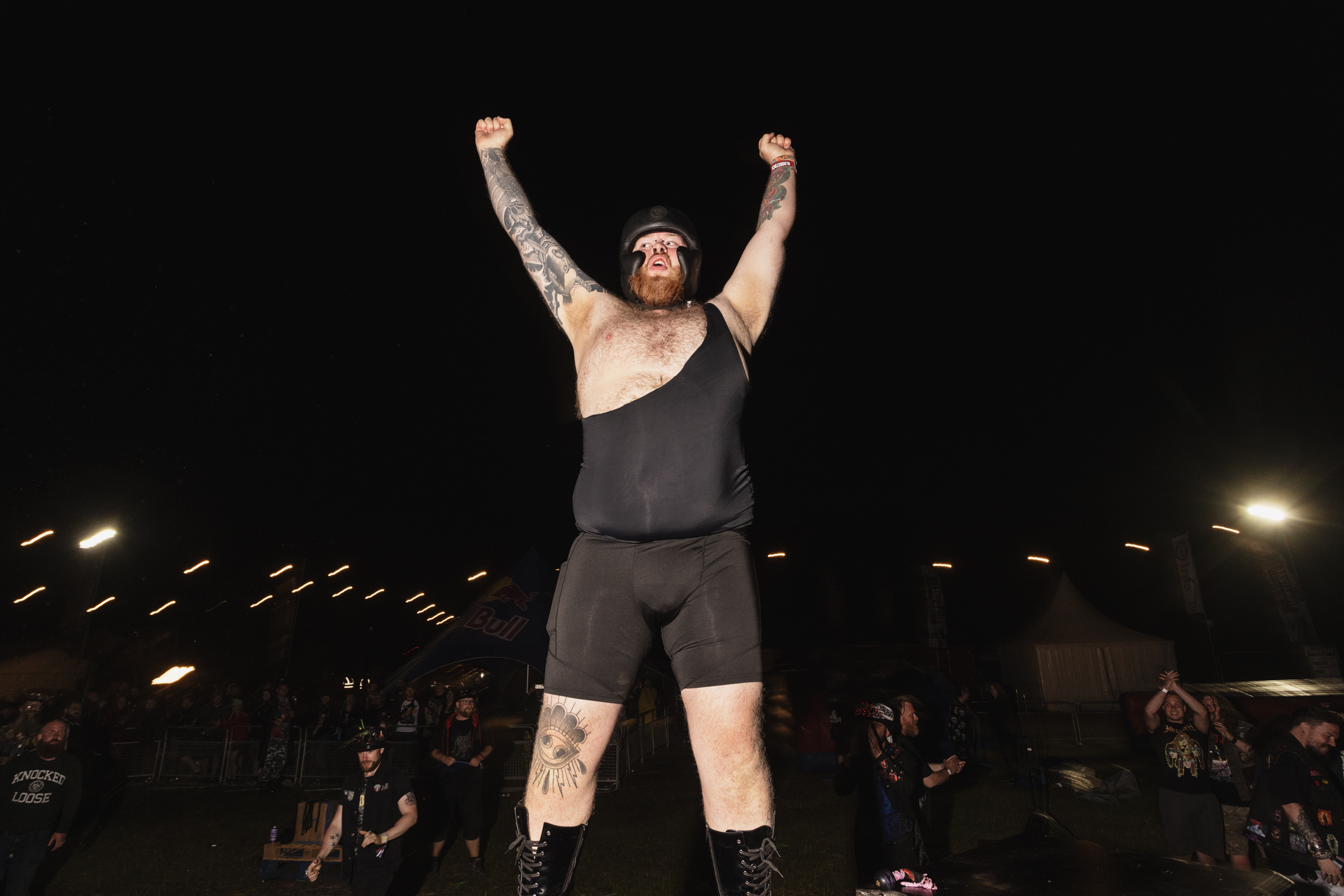 Shirtless wrestler with arms raised in victory pose, wearing black vest and shorts, standing in wrestling ring with crowd in background.
