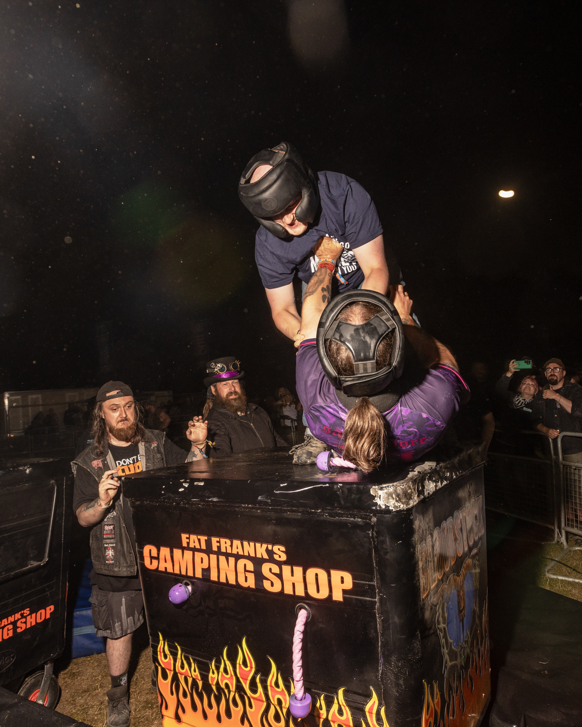Man in grey shirt and cap stands on table branded "Fat Frank's Camping Shop" with flame design, holding large pan over seated audience.